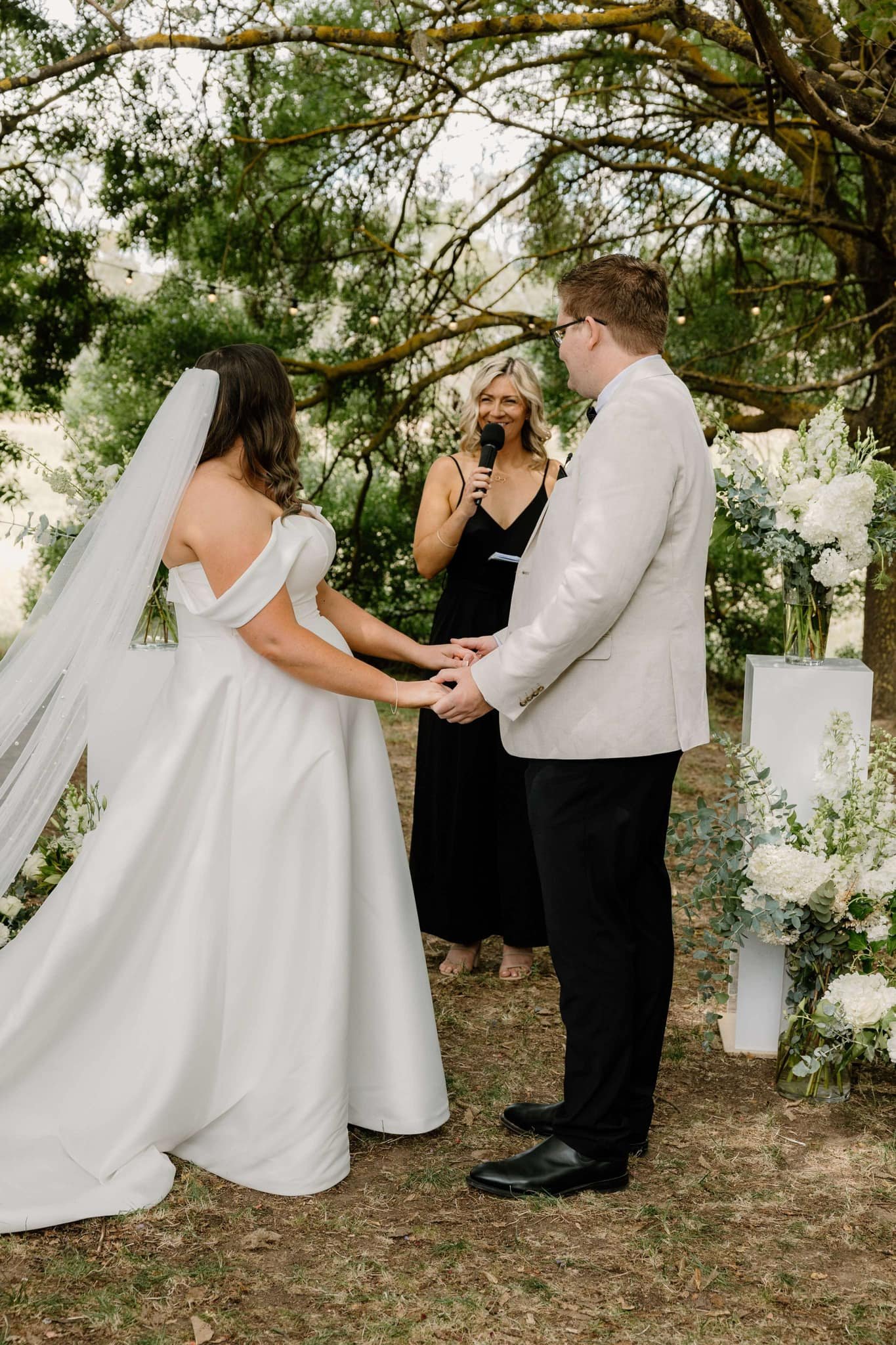 A bride and groom stand holding hands during an outdoor wedding ceremony, with the officiant speaking into a microphone surrounded by greenery and floral arrangements.