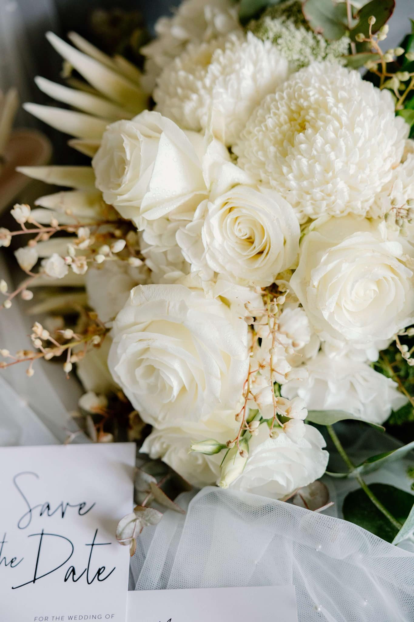 A bouquet of white roses and chrysanthemums with foliage, alongside a "Save the Date" card on a tulle fabric.
