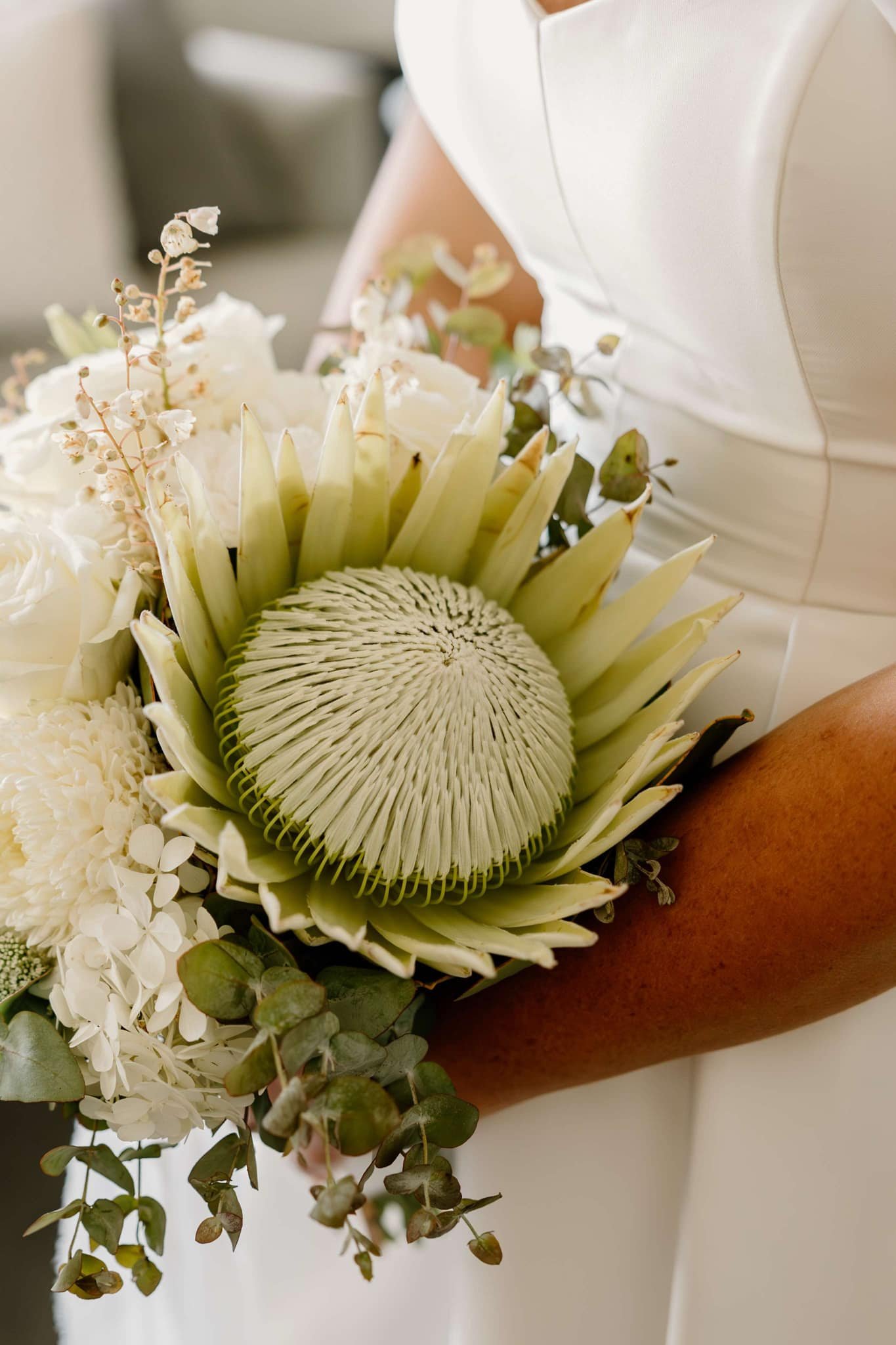 Close-up of a wedding bouquet featuring a large protea flower, white roses, and greenery, held by a bride in a white dress.