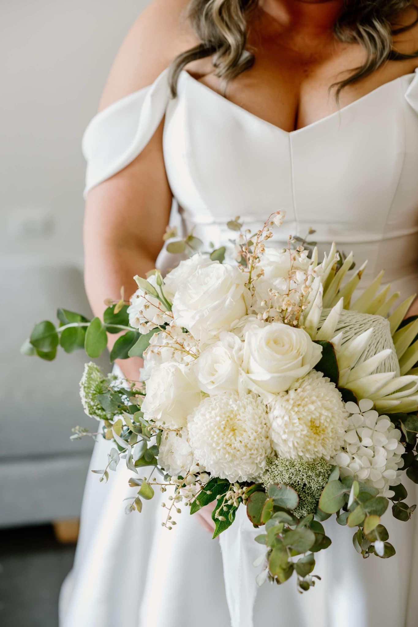 Bride holding a white floral bouquet composed of roses, chrysanthemum, and greenery, wearing an off-shoulder wedding dress.