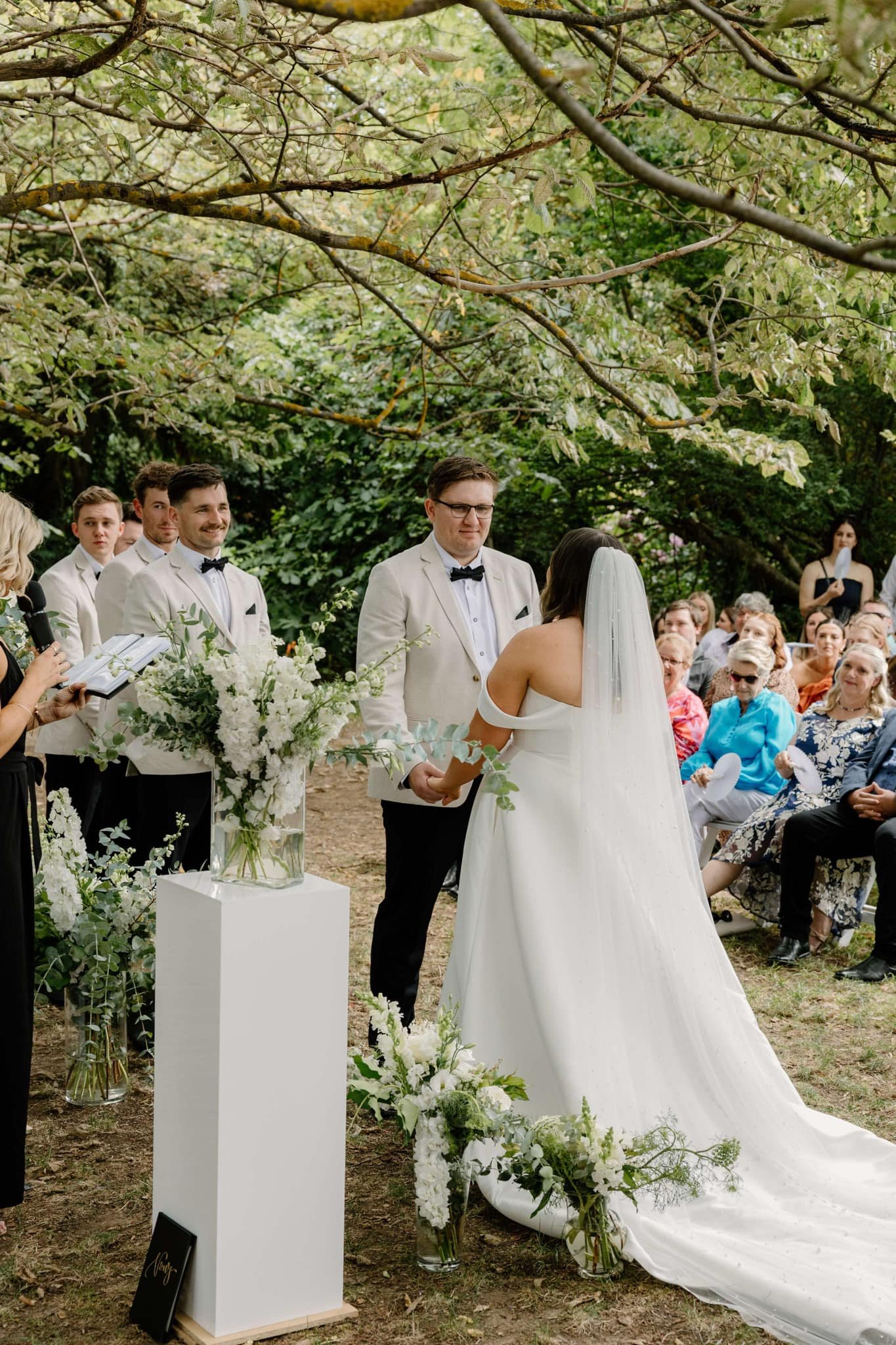Outdoor wedding ceremony under trees with a bride in a white gown and veil, a groom in a white suit, and guests seated. The setting includes floral arrangements and a celebrant holding a book and microphone.