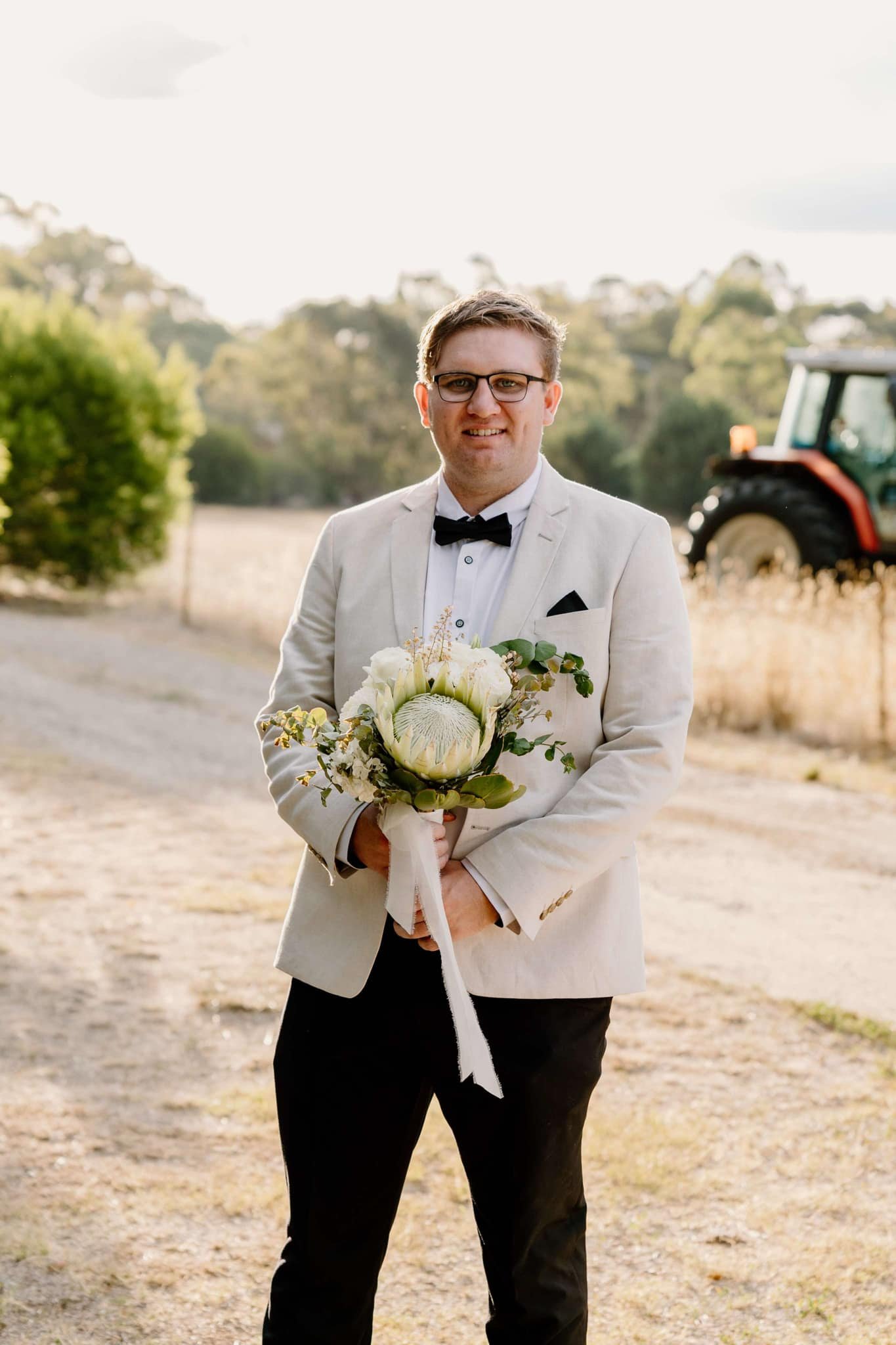 Man holding a bouquet, wearing a light suit with black pants and a bow tie, standing outdoors with a tractor in the background.