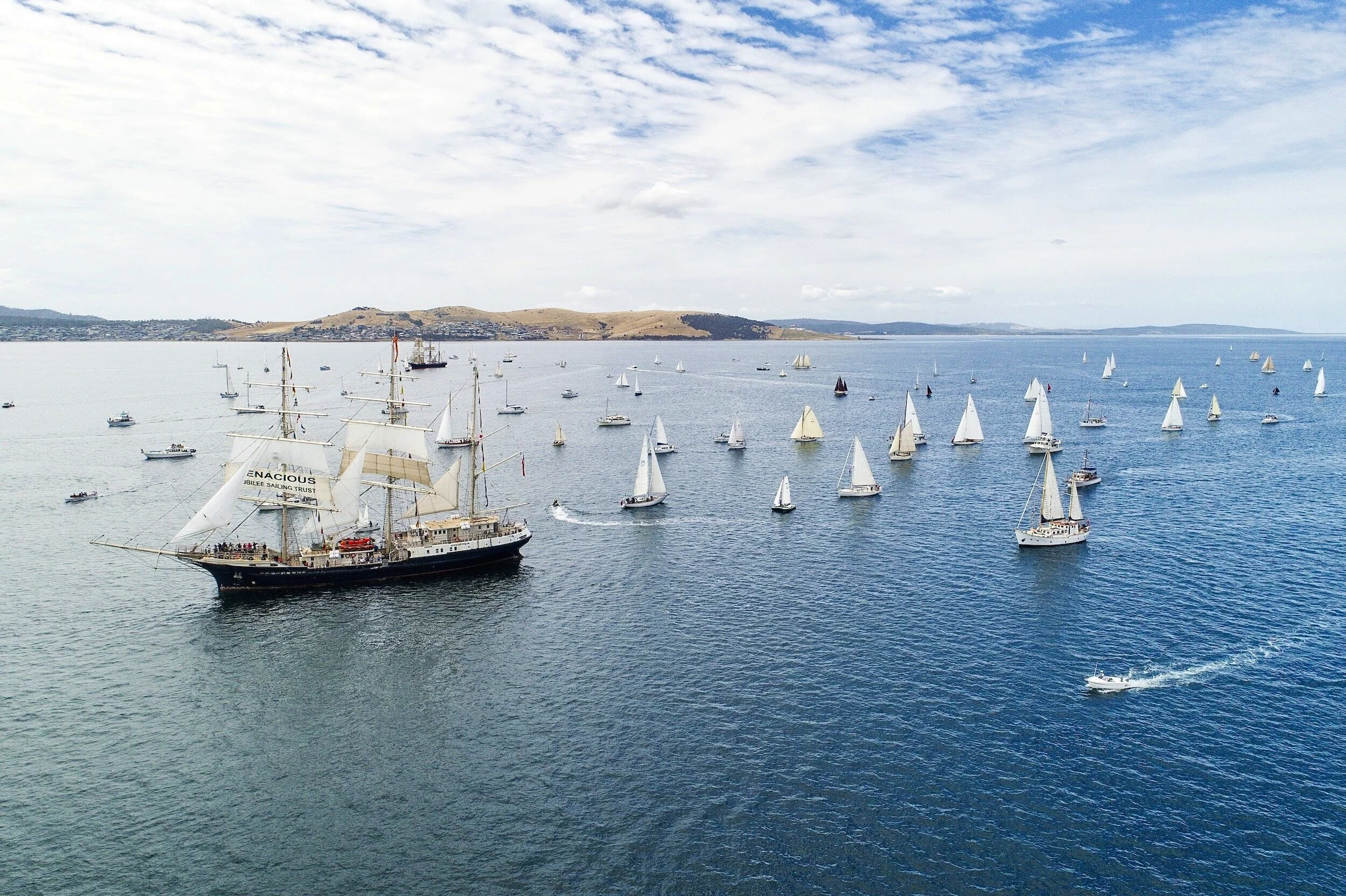 A large sailing ship with multiple masts and white sails, sailing in a body of water surrounded by numerous smaller sailboats and motorboats, with a distant shoreline and hills under a partly cloudy sky.