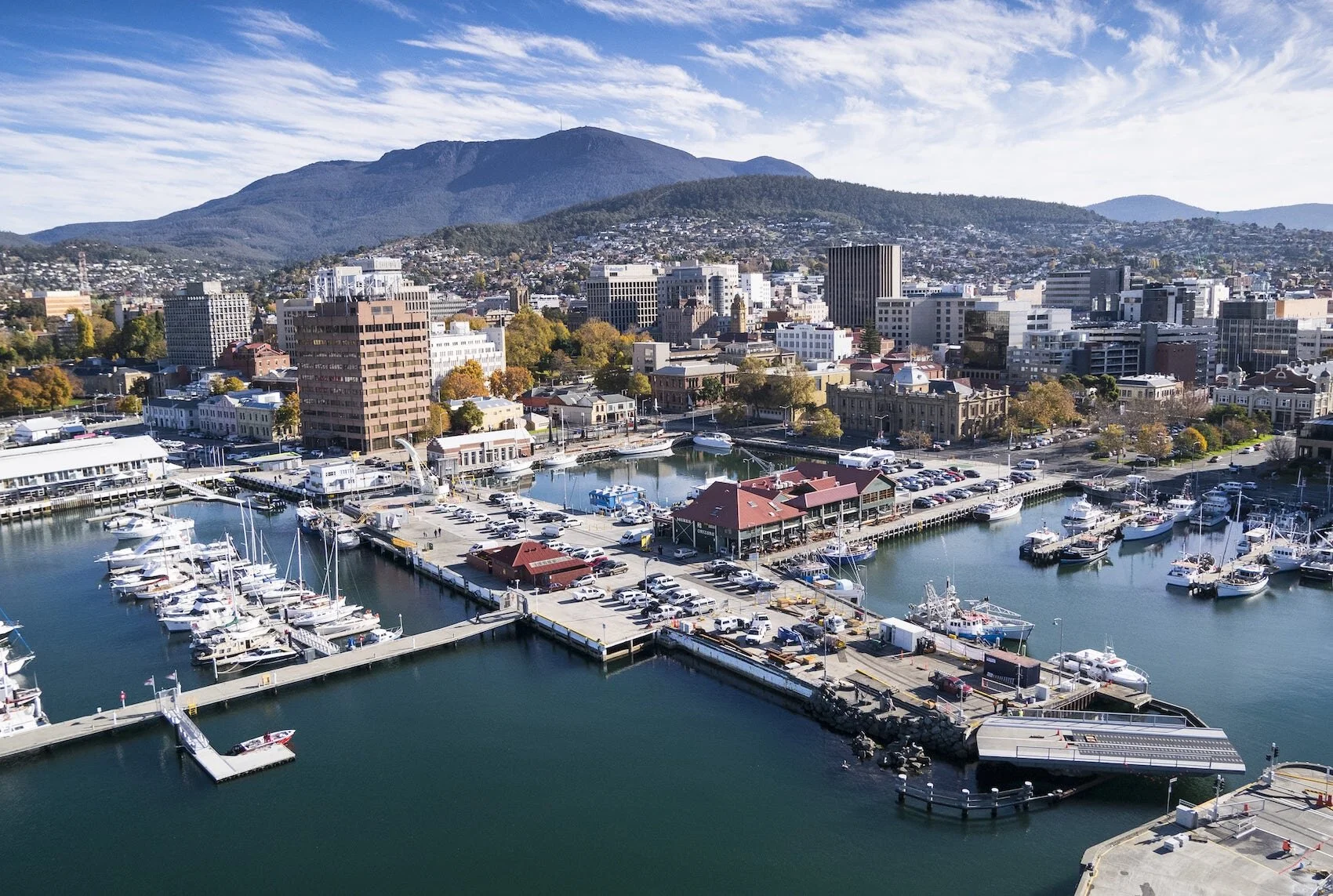 Cityscape of a coastal downtown area with a marina filled with yachts, surrounded by tall buildings, with a mountain in the background under a partly cloudy sky.