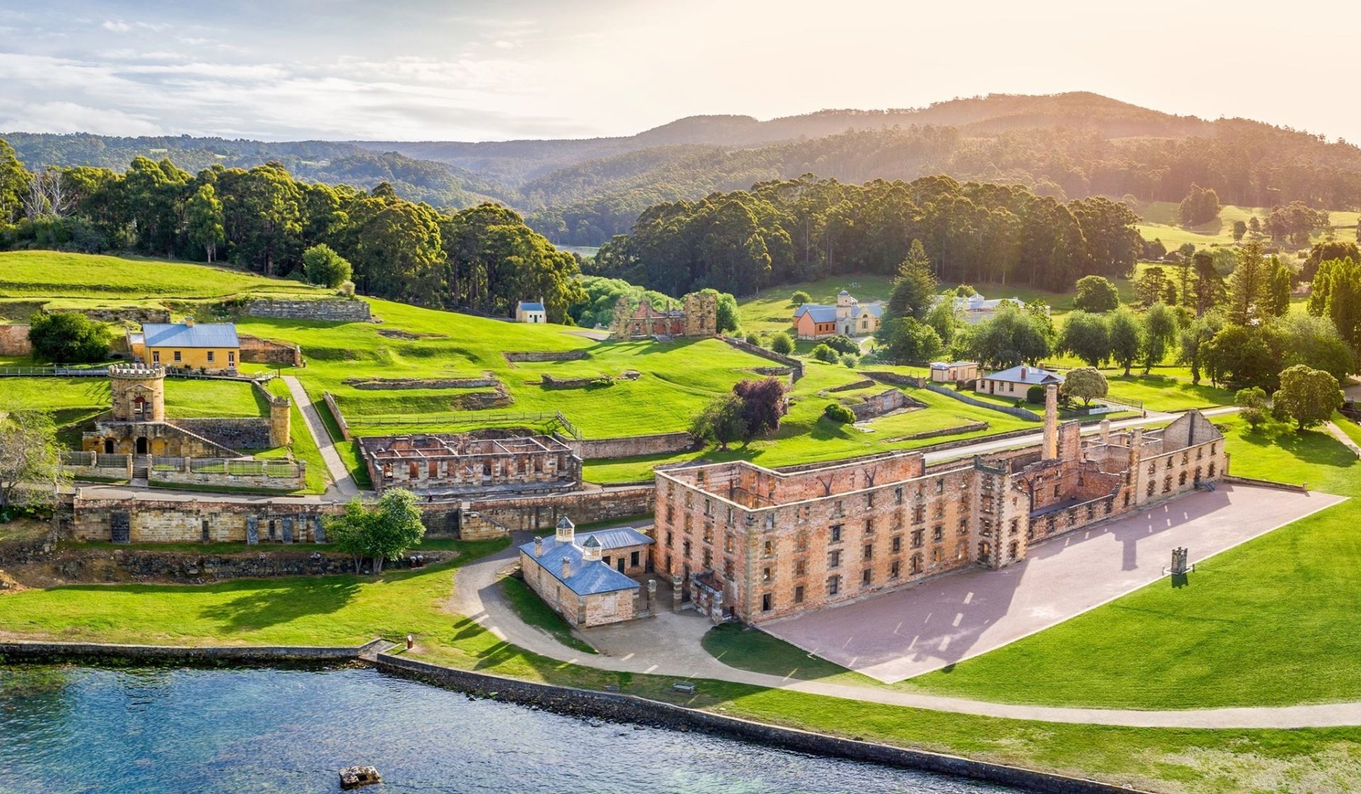 Aerial view of a historic castle surrounded by green lawns, trees, and rolling hills with mountains in the background, during sunset.