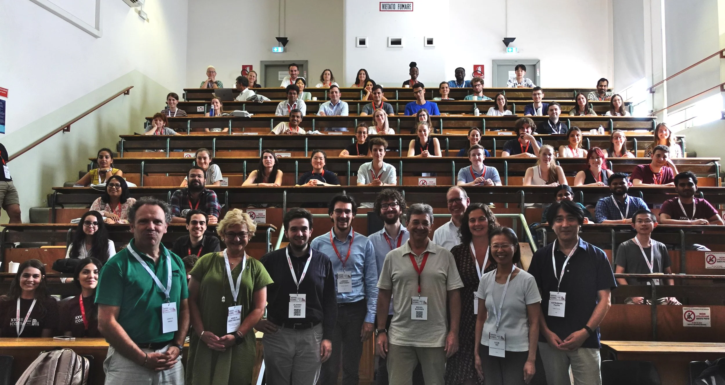 Group photo of many senior and early career researchers in a lecture hall at a summer training school, wearing conference badges.