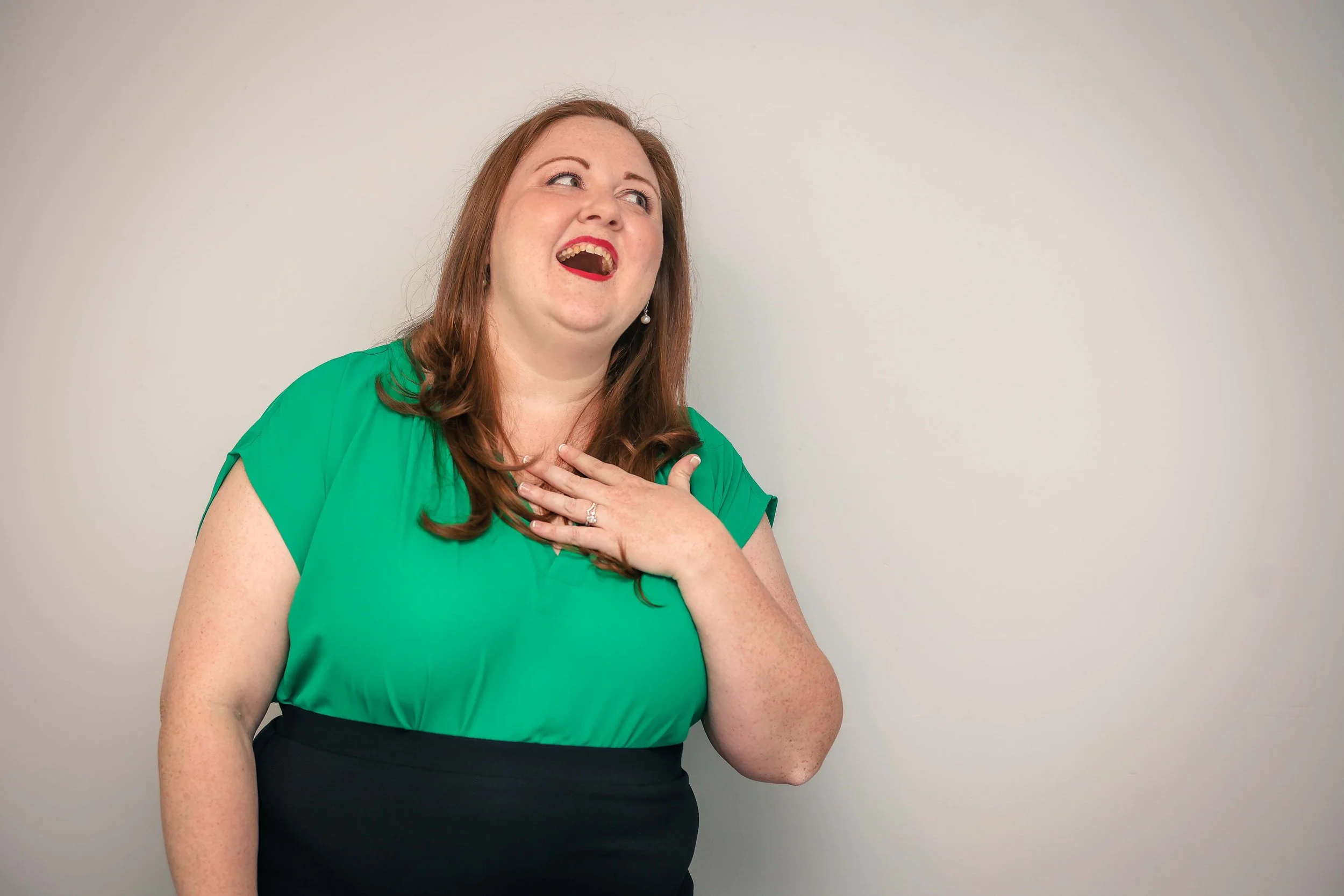 A woman wearing a green top and black skirt, with red hair, smiling with her hand on her chest, standing in front of a plain light-colored wall.