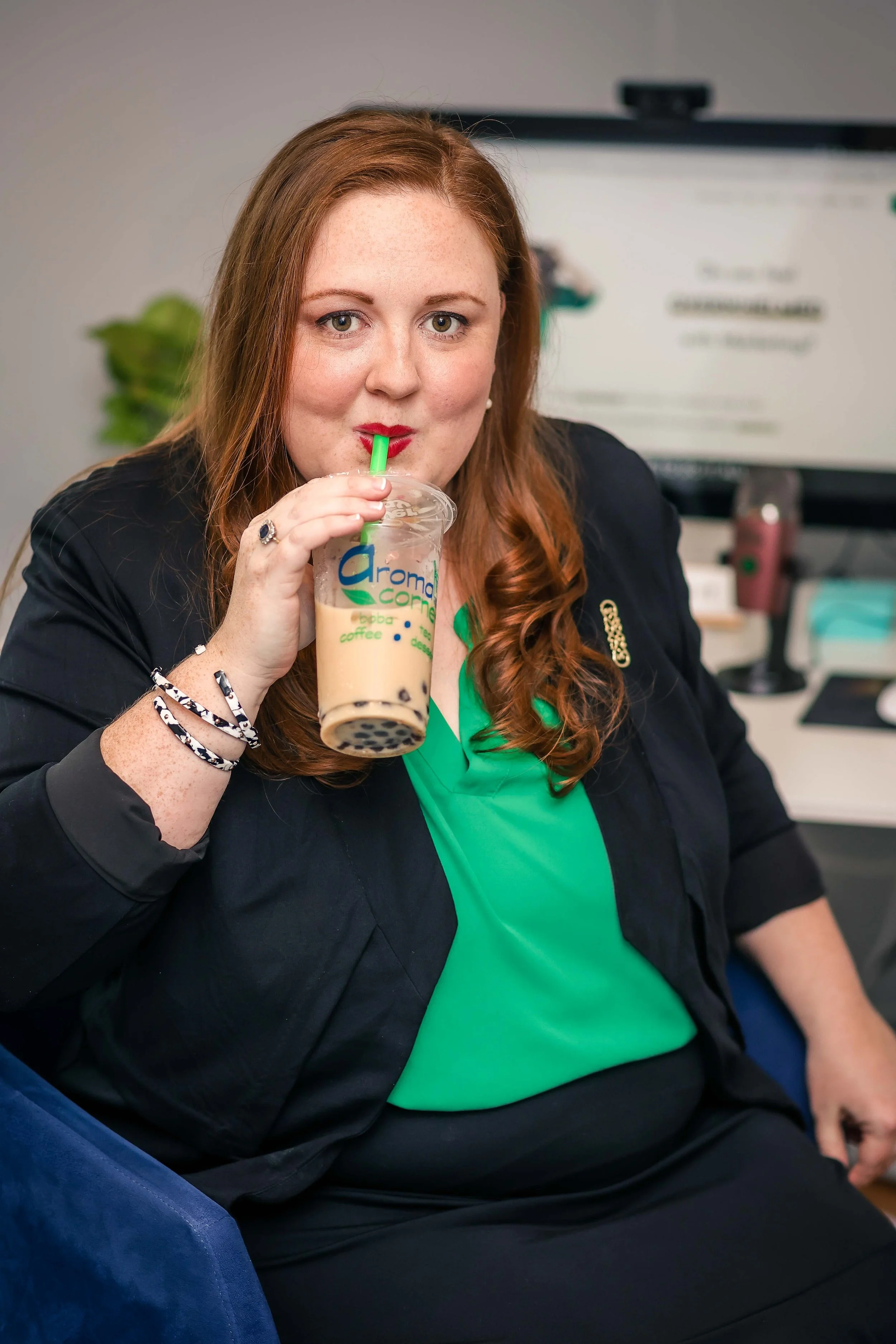 A woman with long red hair, wearing a black jacket and a green shirt, is sitting in a chair and drinking bubble tea through a green straw.