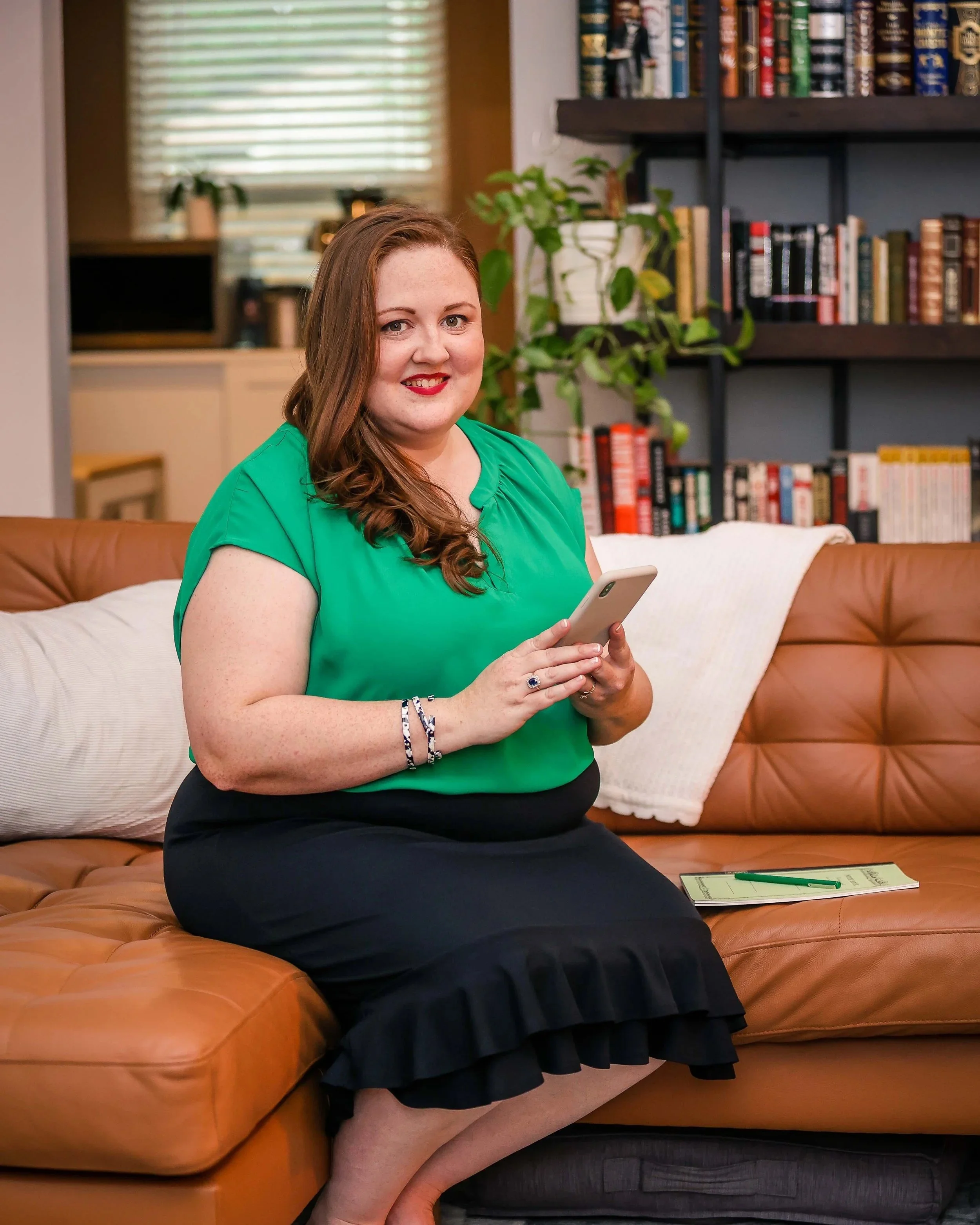 Woman with light skin and long wavy brown hair sitting on a tan leather couch, holding a smartphone, wearing a green blouse and a black skirt. Behind her are bookshelves and a window with blinds, a potted plant, and a white blanket on the couch.