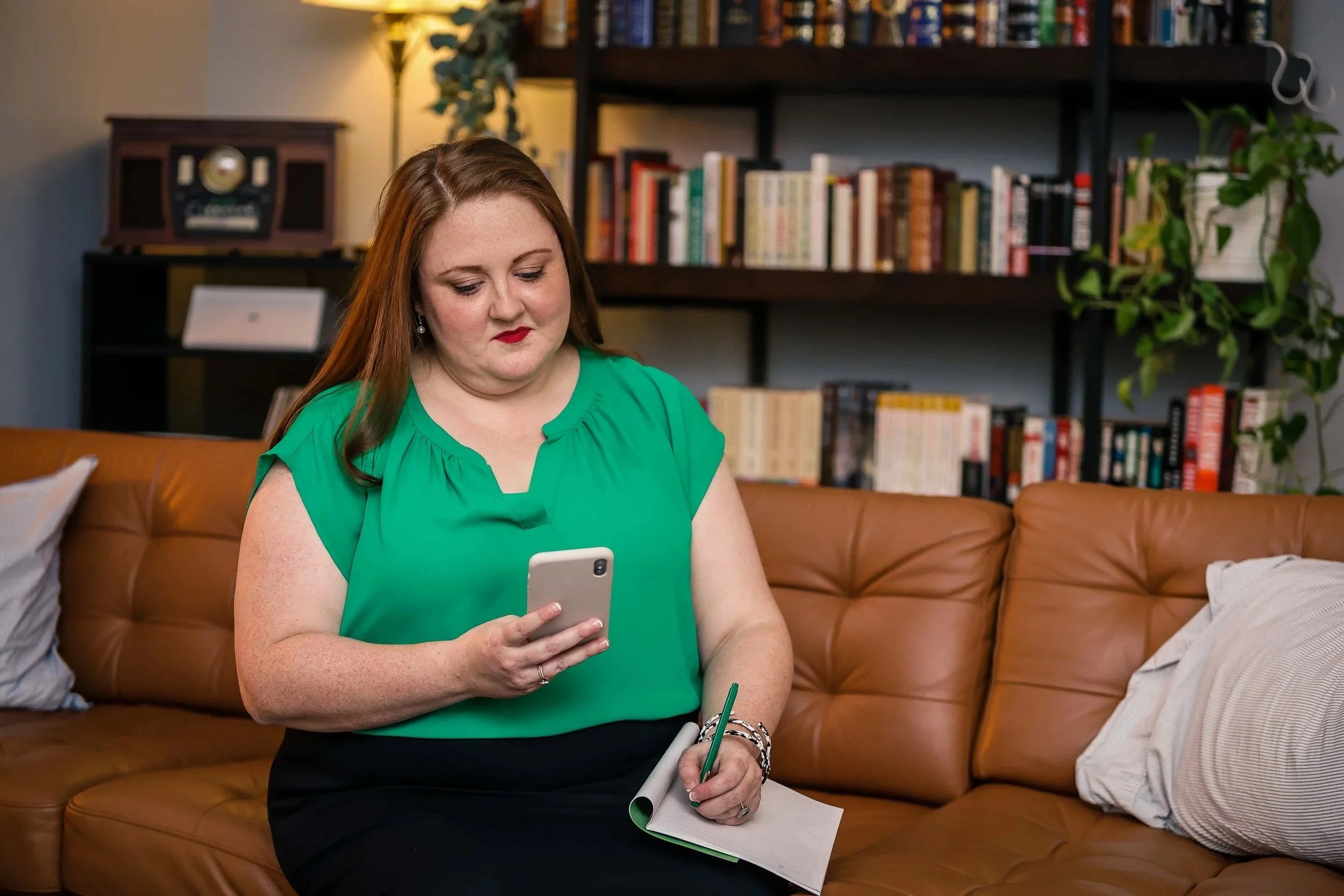 Woman in green blouse sitting on a brown leather couch, looking at her smartphone, with a notebook and pen in her lap, in a room with a bookshelf and plants.