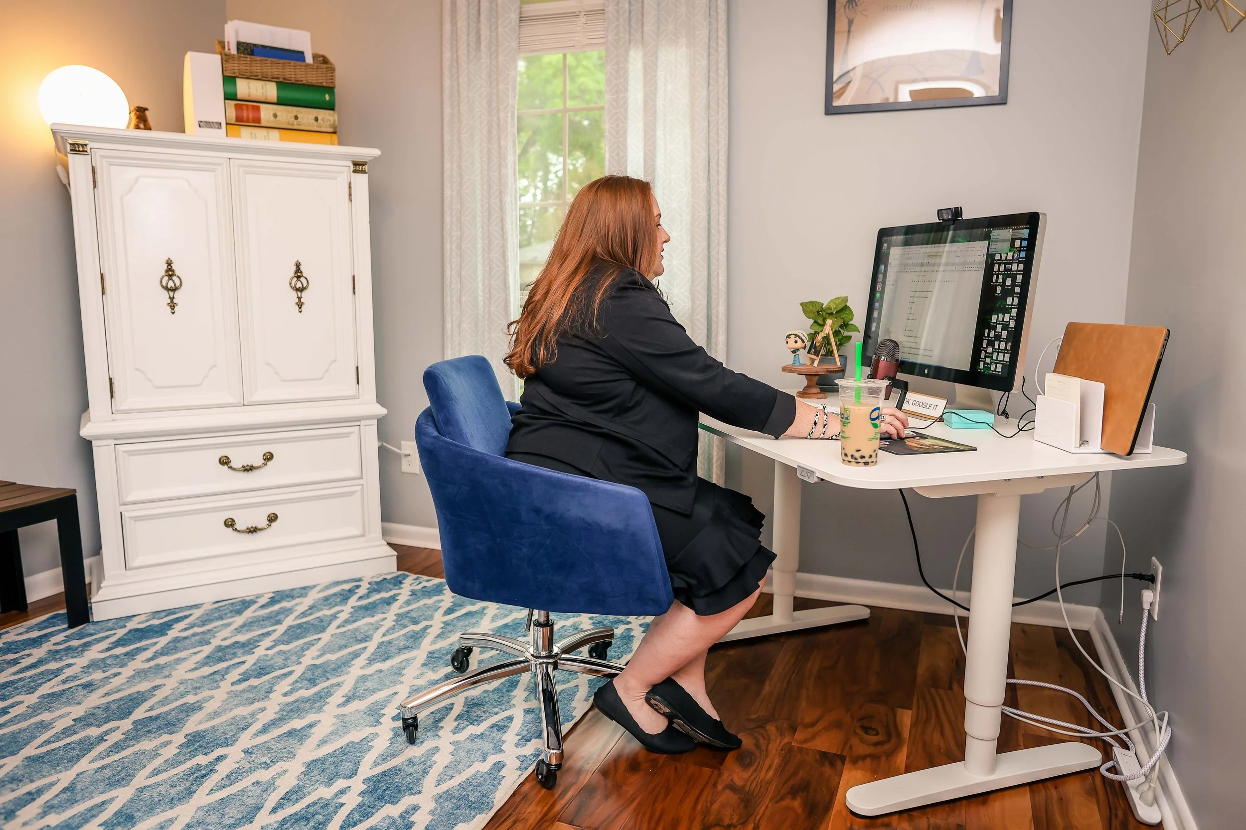 A woman with red hair sitting at a white desk using a computer in a home office. She has a bubble tea drink on her right and a small plant and figurine on her left. Behind her is a window with sheer curtains, a white cabinet, a framed picture, and some hanging decorative objects.