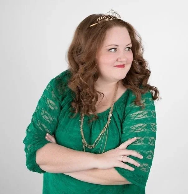 A young woman with curly red hair wearing a green lace top, a tiara, and layered necklaces, posing with arms crossed and looking to the side against a plain light background.