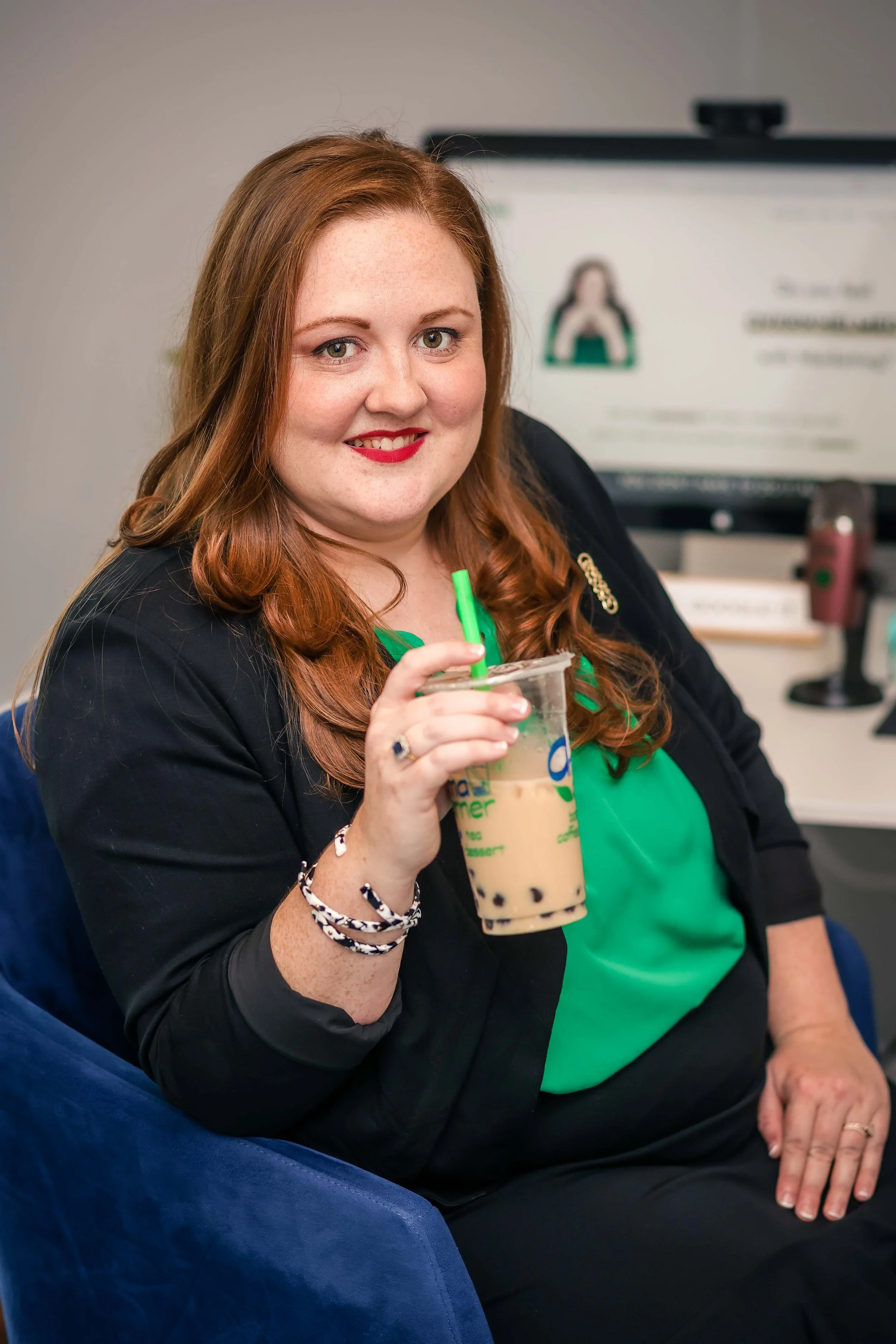 A woman with long red hair and fair skin, smiling and holding a large iced coffee drink with black tapioca pearls, sitting in a chair in front of a whiteboard, wearing a black jacket and a green shirt.