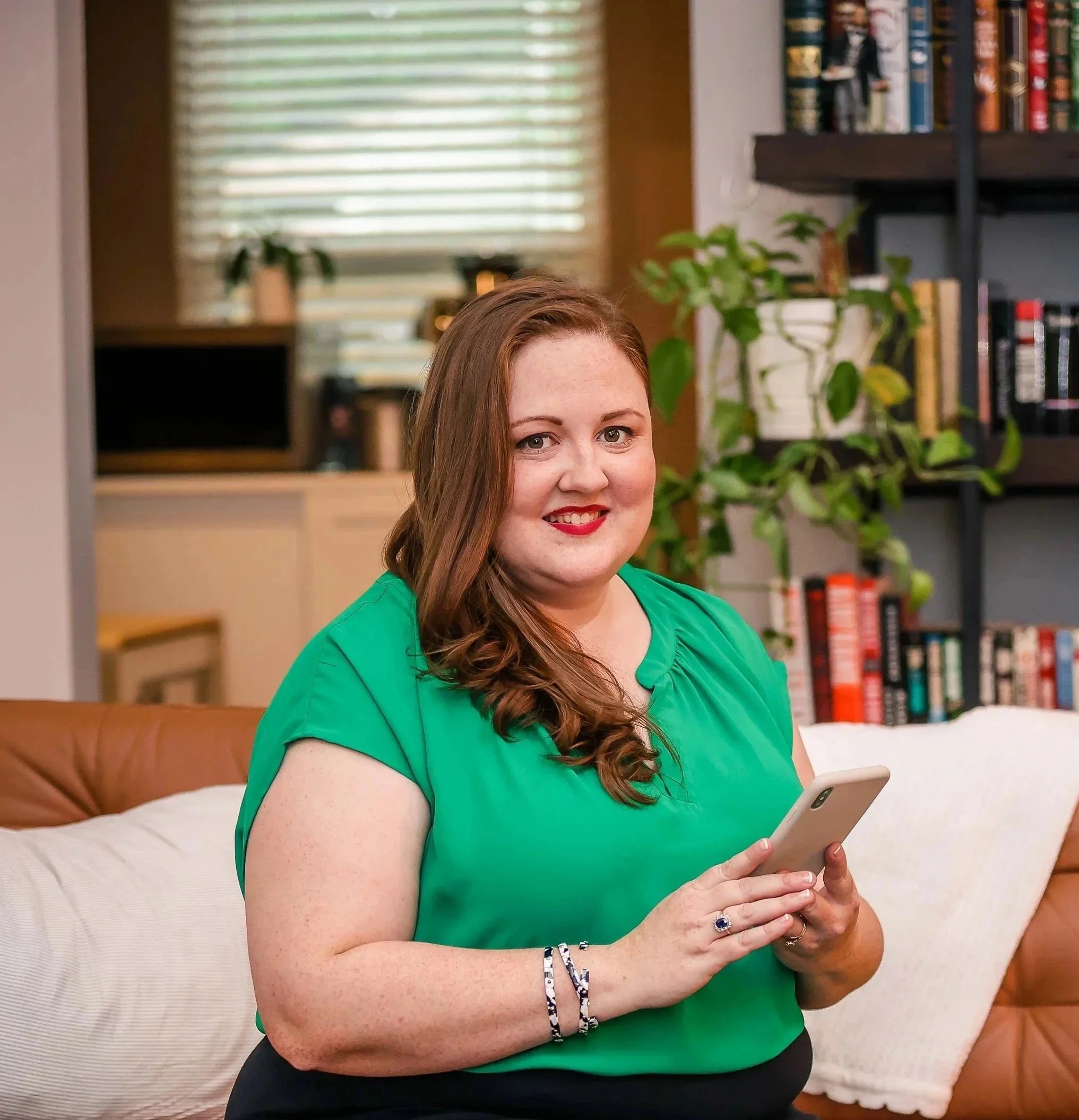 A woman with long red hair and a bright green shirt sitting indoors, holding a smartphone. She is wearing rings and bracelets, and has a friendly expression. In the background, there are bookshelves with various books and some indoor plants.