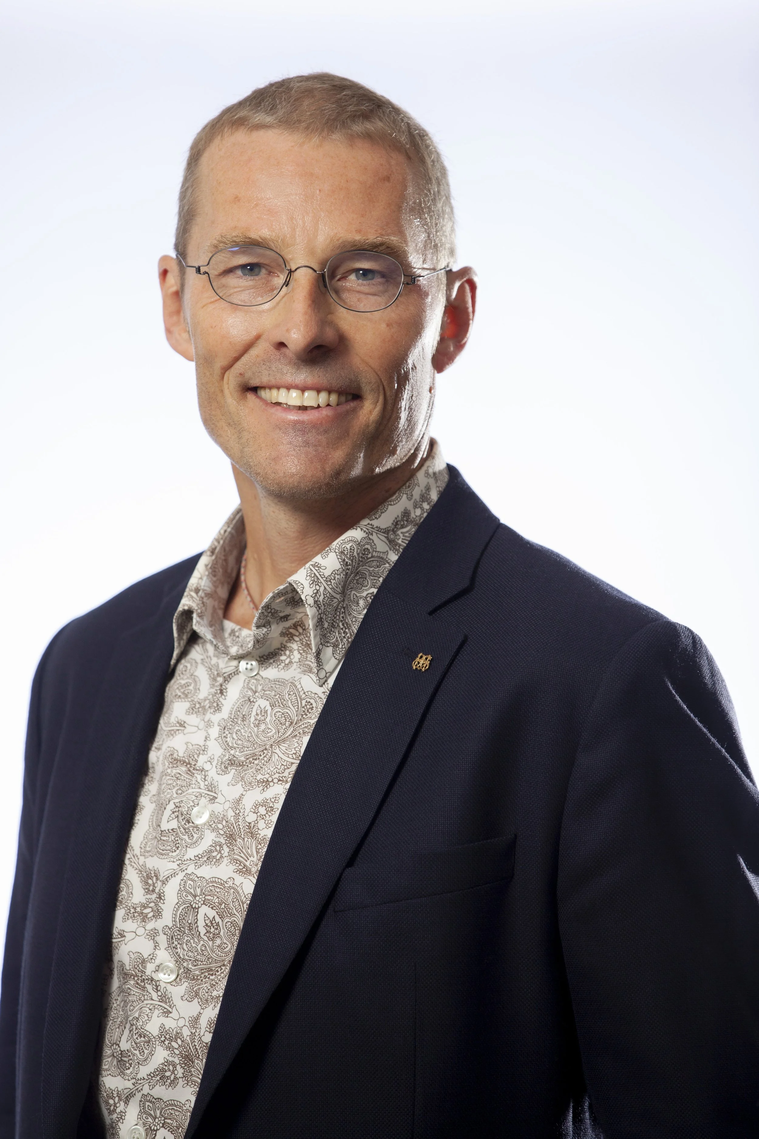 Man i mörkblå kostym och vit blus med paisleymönster, ler och bär glasögon, mot vit bakgrund.