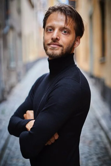 En man med svart tröja står med korsade armar på en gata med kullerstensbeläggning.
