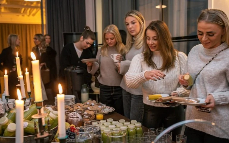 Grupp av unga kvinnor som väljer mat vid buffébord med bakverk, frukt och drycker, i ett rum med moderna fönster och ljus stämning.