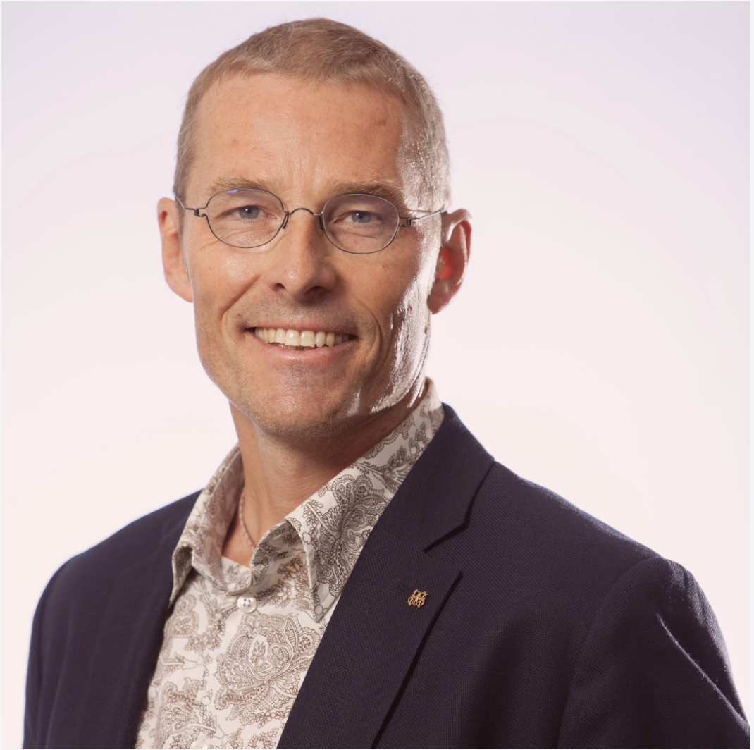 Porträtt av en man med kort, ljus hår, glasögon och ett leende, klädd i en mörk kavaj och en mönstrad skjorta, mot en vit bakgrund.