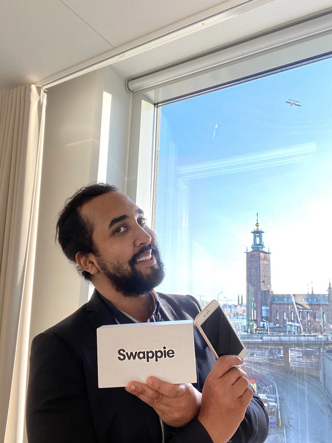 Man med mörk hår och skägg, skrattar och håller ett kort med 'Swappie' framför sig, står vid ett fönster med en stadsskyline och ett flygplan på himlen.