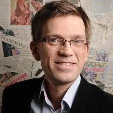 Man i kavaj och lätt blå skjorta med gläsögon, mot en bakgrund av vägg täckt av tidningsurklipp och papper.