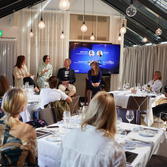 En paneldiskussion med fem personer framför en publik, med en stor skärm bakom dem. Publiken äter vid dukade bord i ett elegant konferensrum.