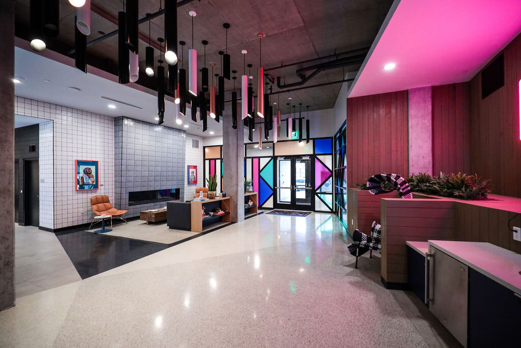 An interior image of the Neon Local Lobby illuminated with pink neon lights. Includes a minimalistic stained glass window in the entry and various lights hanging from the ceiling. The walls include a combination of tile, wood, and concrete