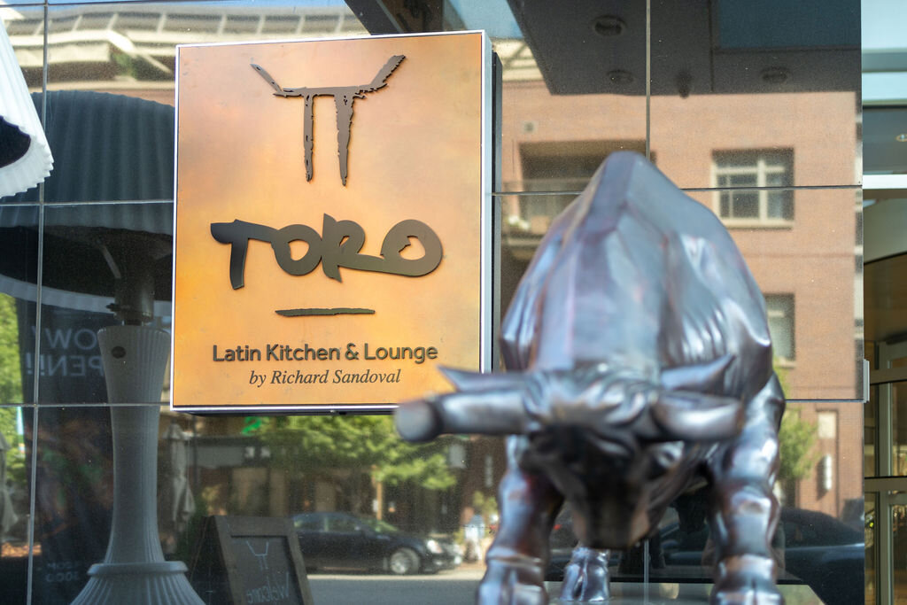 Toro Latin Kitchen and Lounge sign behind a bull statue is an exterior sign that reads, "Toro Latin Kitchen & Lounge by Richard Sandoval"