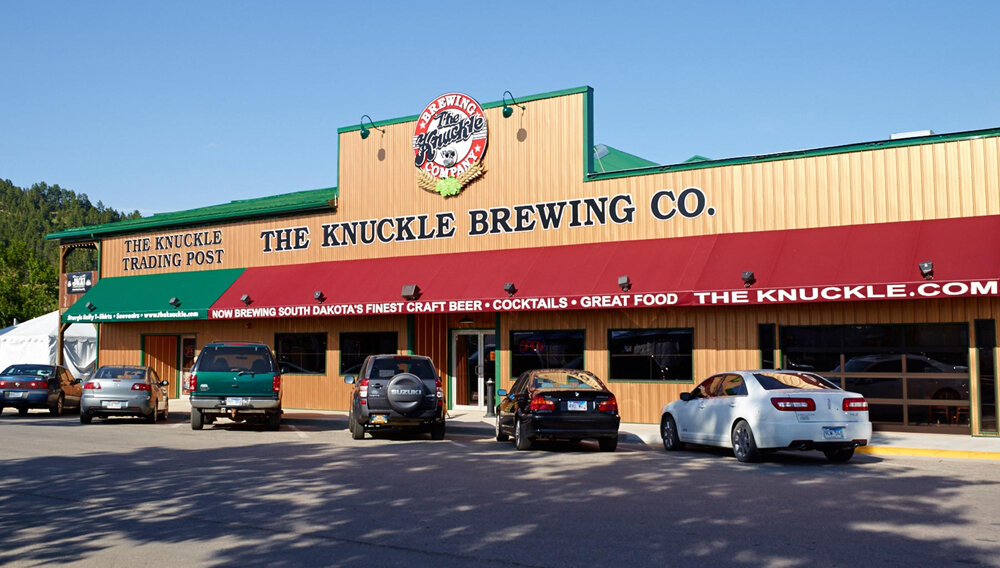 Exterior Image of "The Knuckle Brewing Company" and "The Knuckle Trading Post"" building. Designed like an old western style building with a red awning below the Knuckle Brewing Company and a Green awning below the Trading Post
