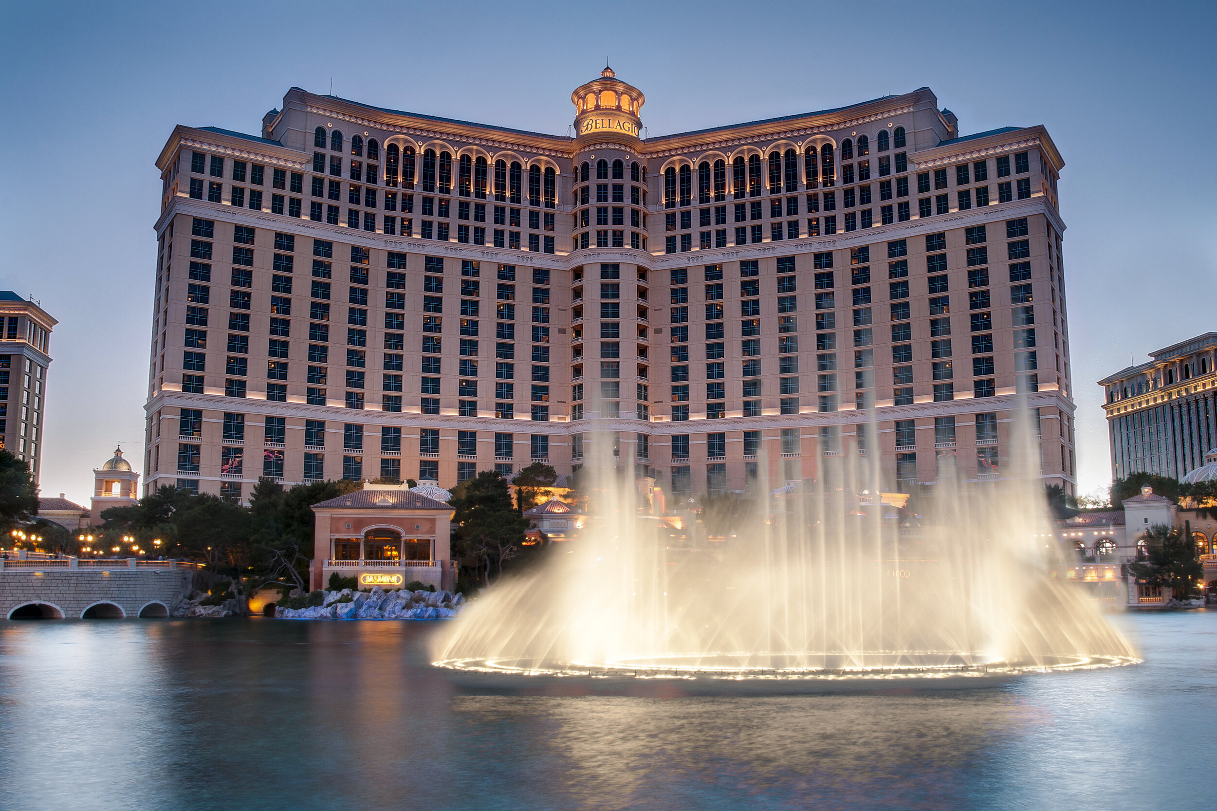 An Exterior image of the Bellagio Hotel with fountains in front. The image is taken in the late afternoon as some building lights are starting to turn on.