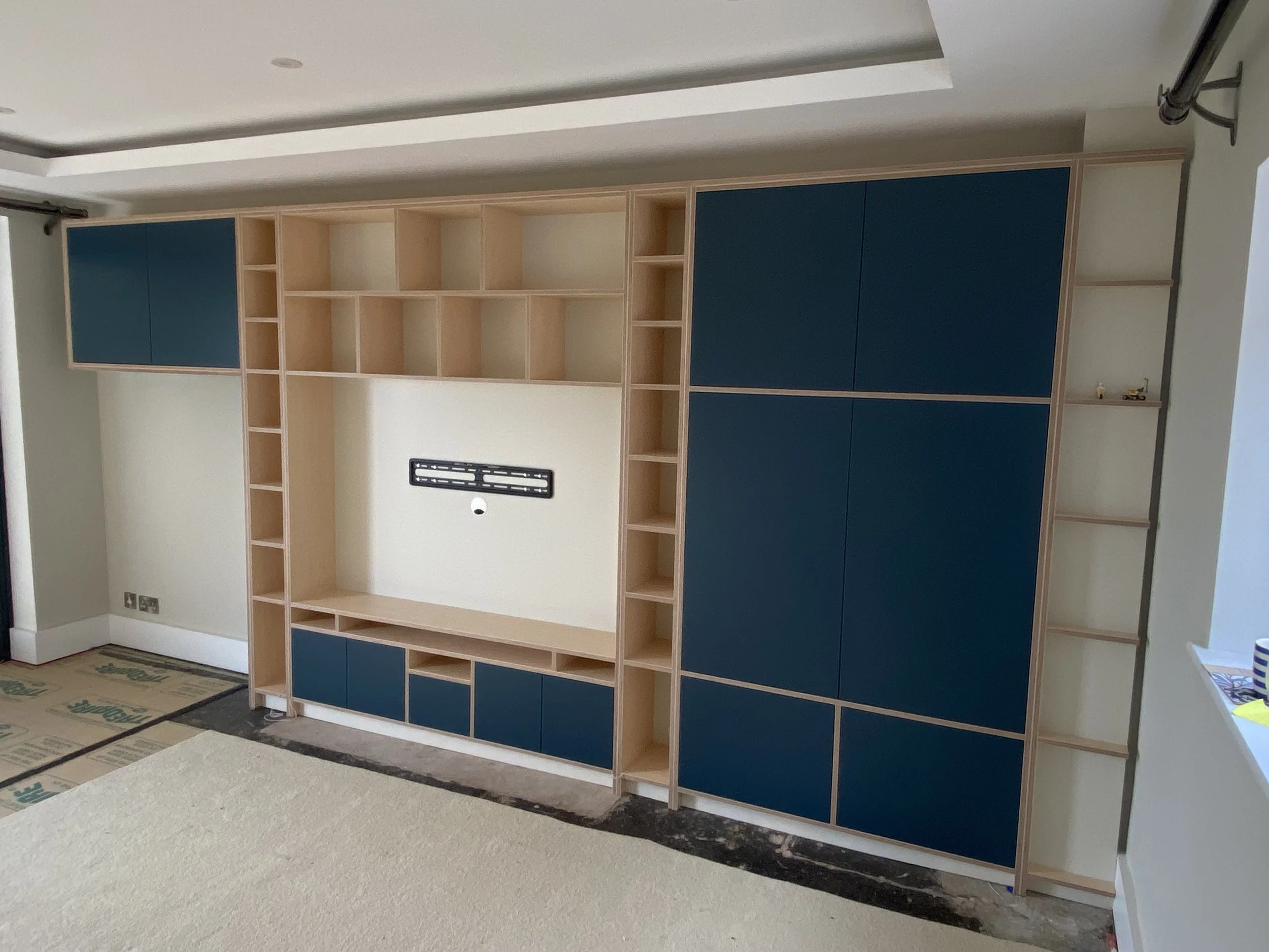 An indoor room or office under construction with a large hybrid shelving unit, part wood and part blue panels, against the wall. The floor is unfinished, with visible insulation and construction materials.