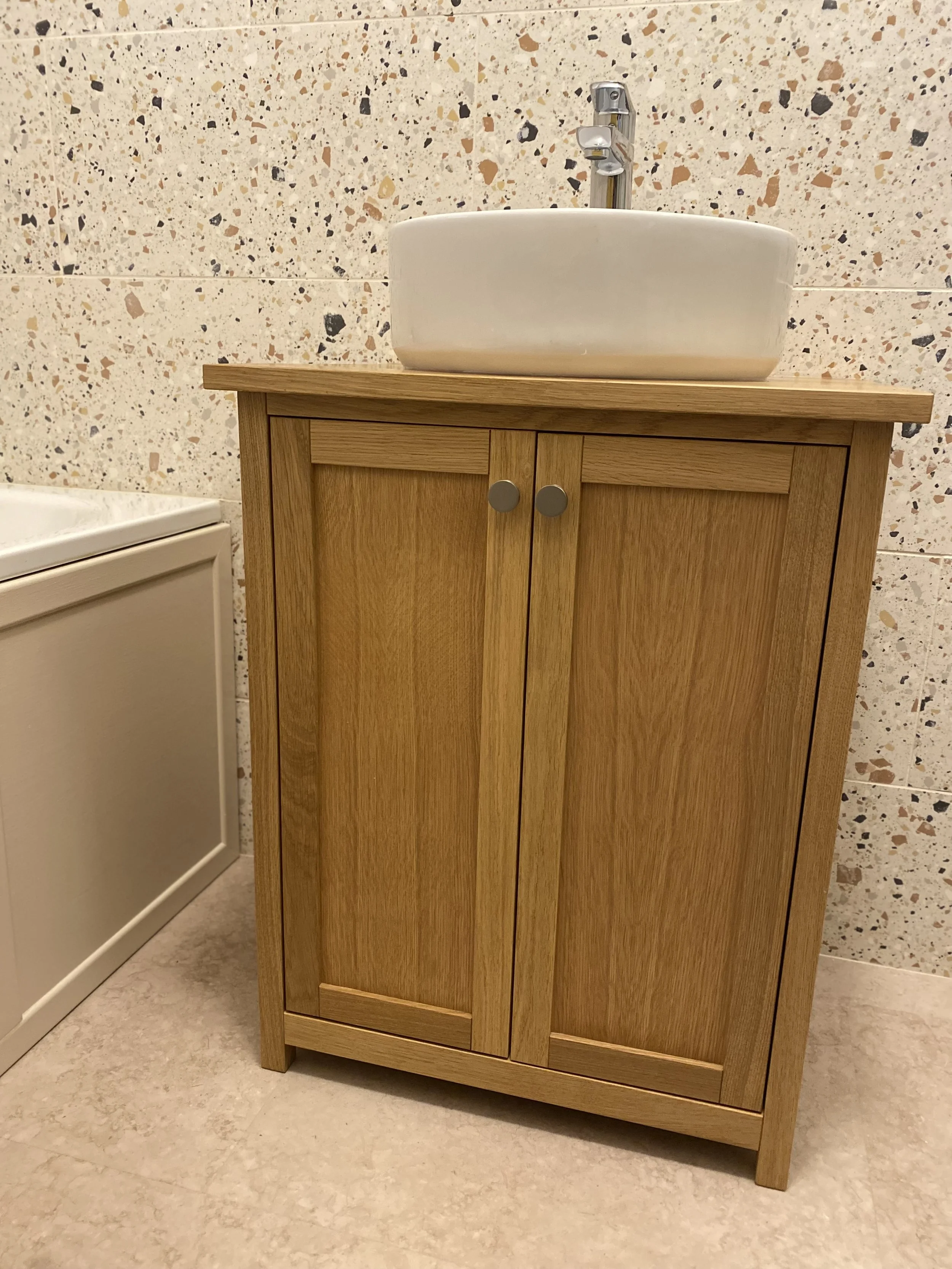 A wooden cabinet with a white ceramic vessel sink on top and a chrome faucet in a bathroom with a speckled tile wall.