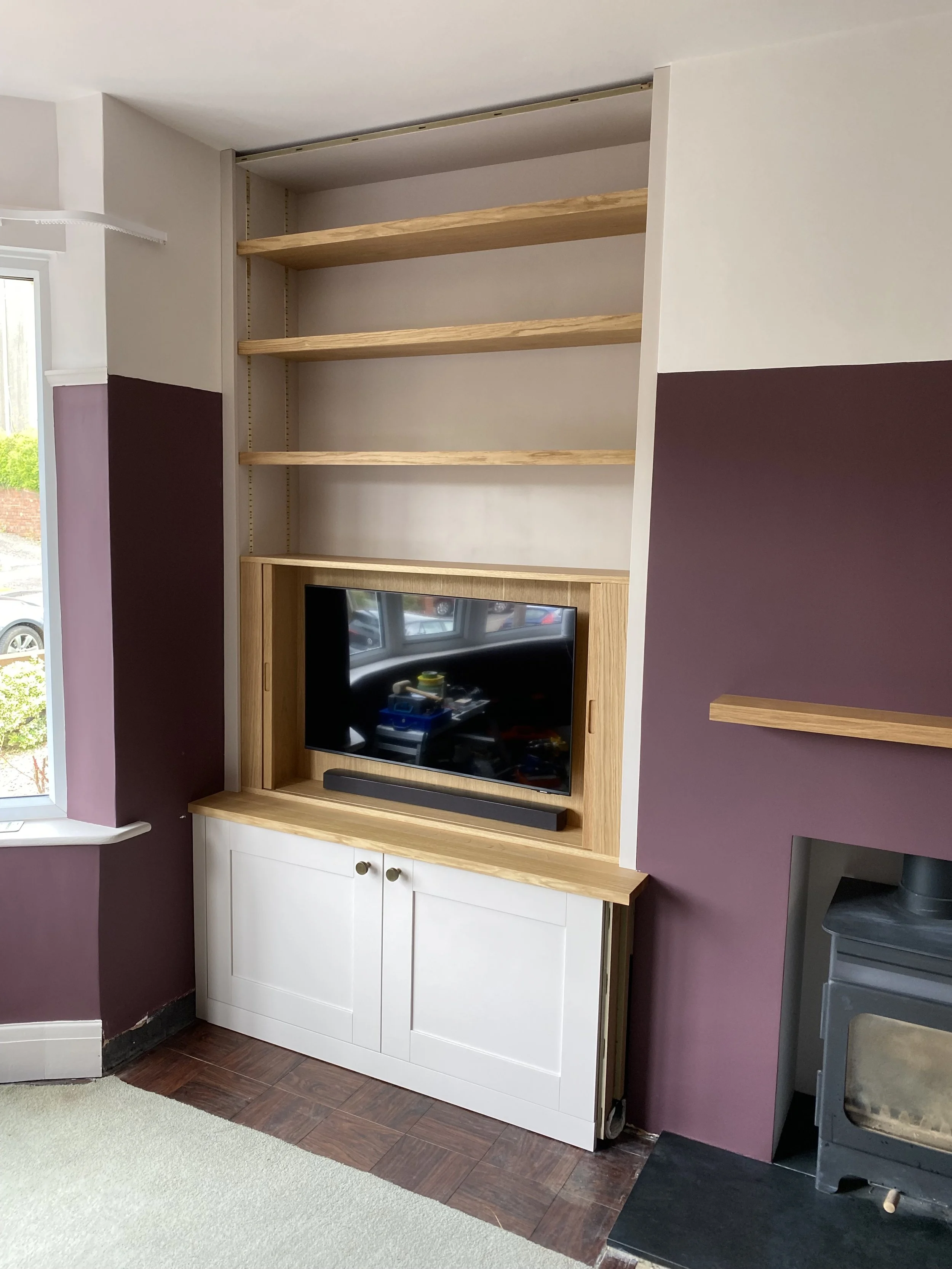 A living room corner with a purple and white built-in shelf unit. The unit has three wooden shelves and a lower white cabinet. A television is placed on the cabinet. To the right, there is a black stove or heater. A large window on the left lets in n