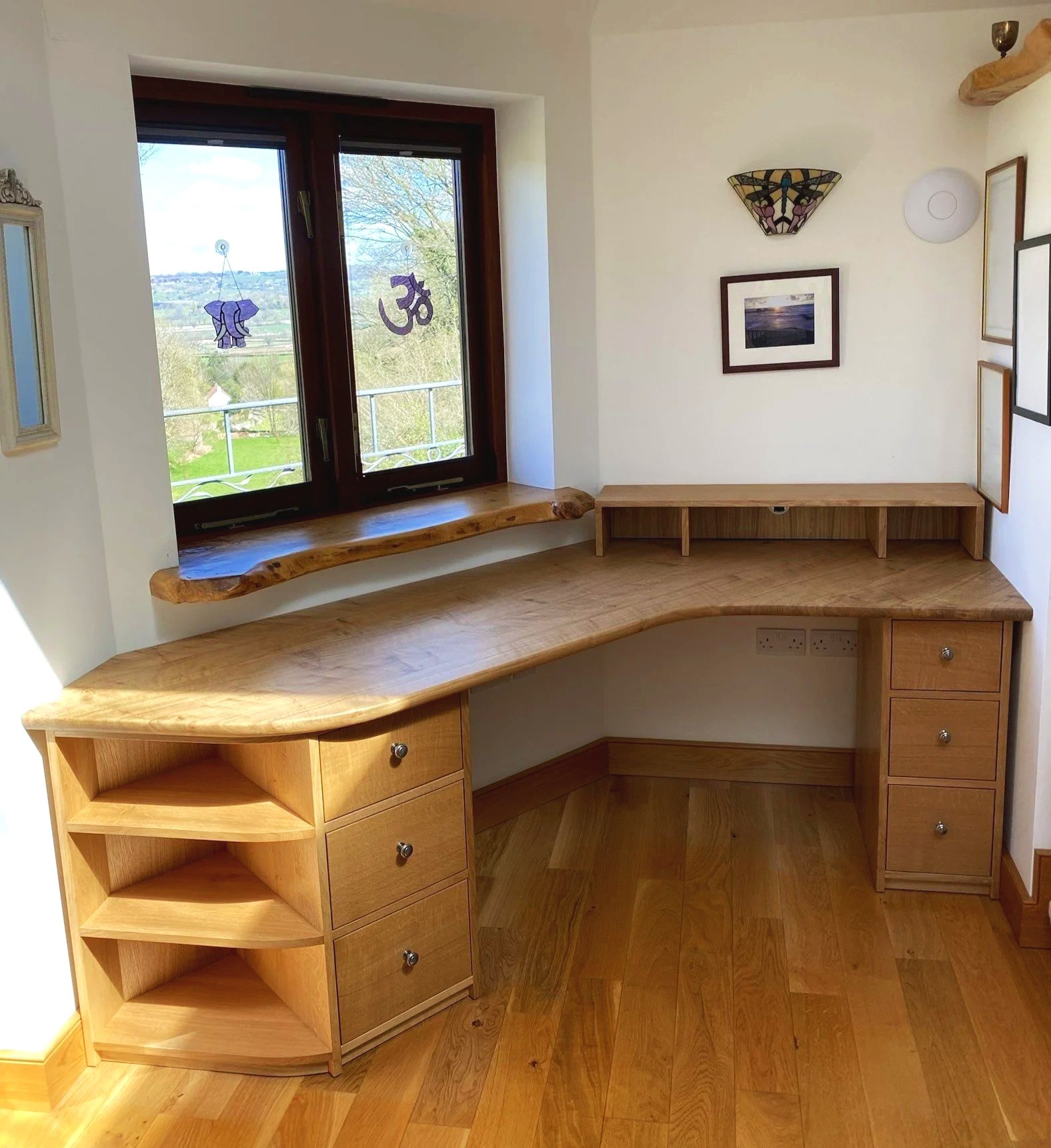 Wood computer desk built around a window with a view of trees and hills, with small shelves and drawers on the left and right sides.