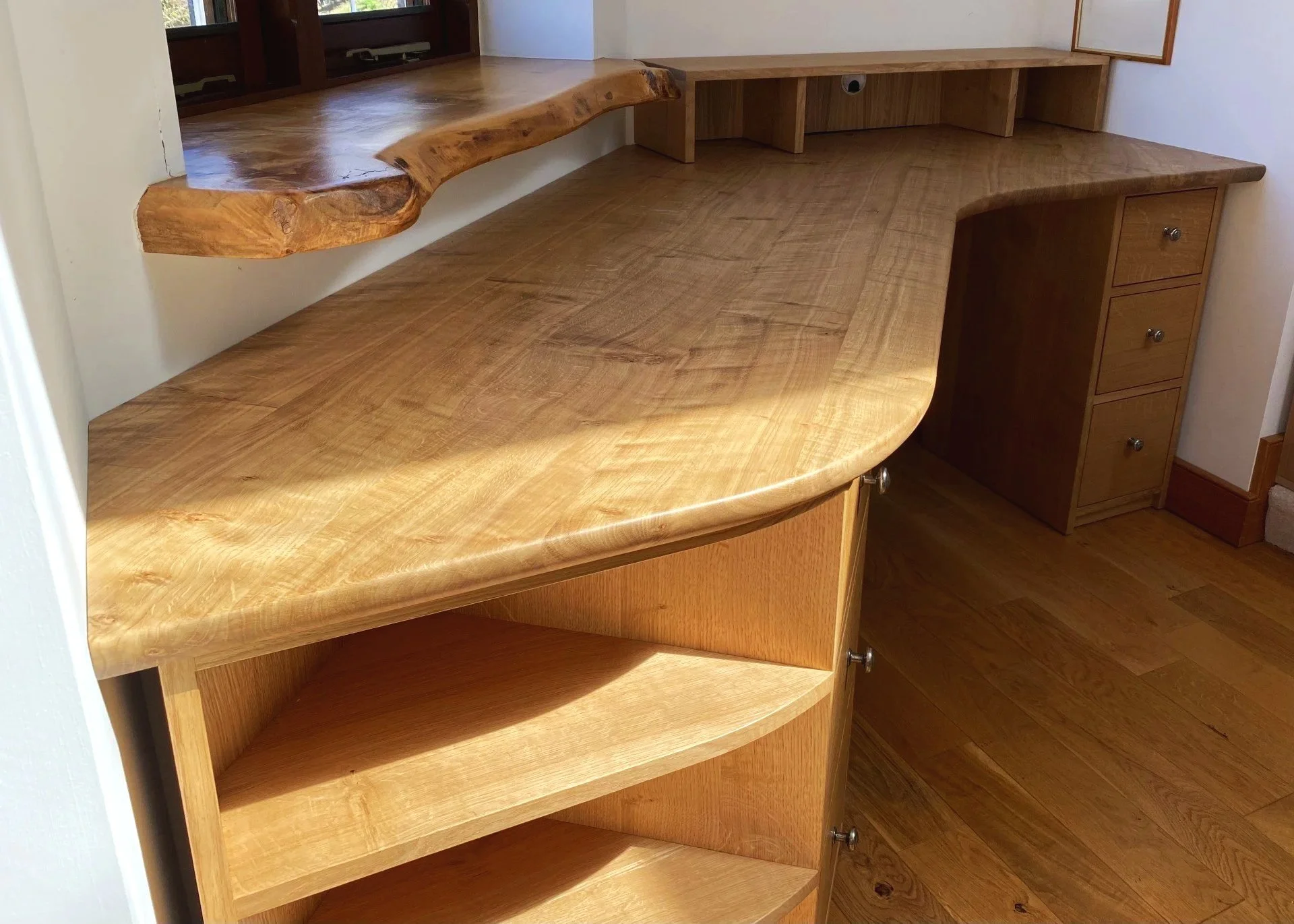 Wooden built-in desk with a live edge top, drawers, and open shelves, situated in front of a window with a wooden frame.