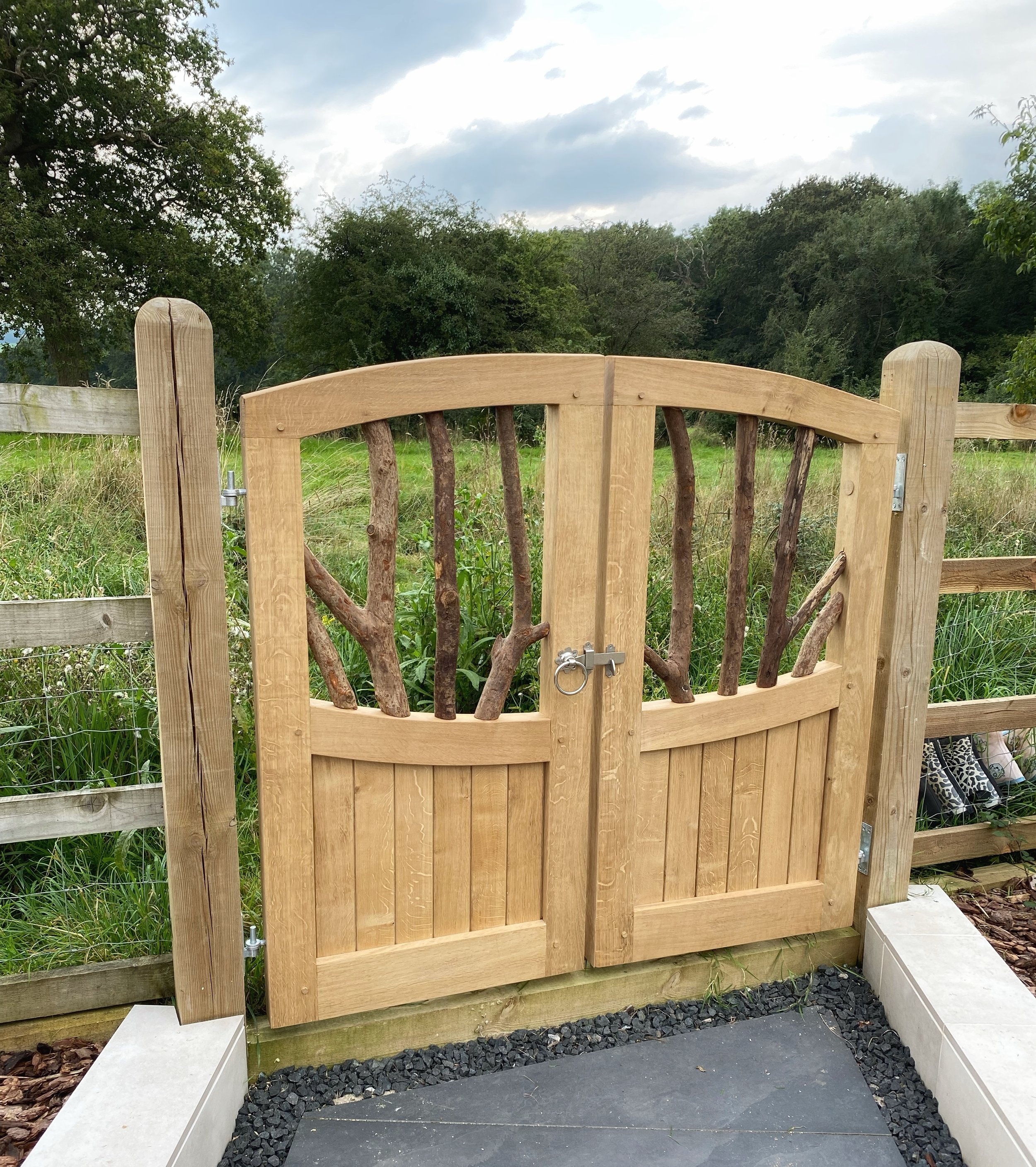 A newly installed wooden gate with vertical and curved horizontal wooden panels, secured with a metal latch, in a backyard with a grassy field, green trees, and a partly cloudy sky in the background.