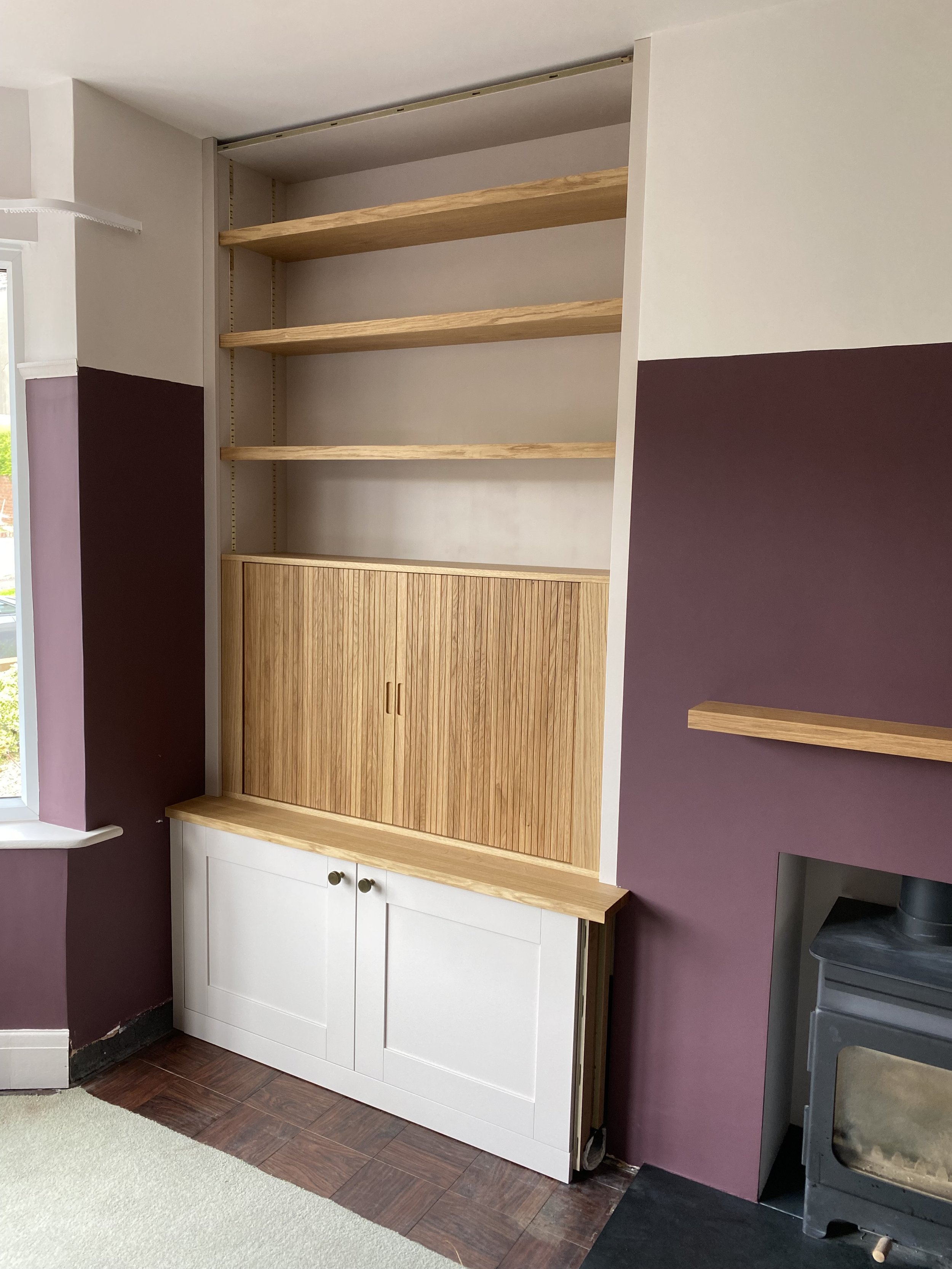 Empty wooden wall shelves and a cabinet in a room with purple and beige walls, partially framed by a corner window and a stove on the right.