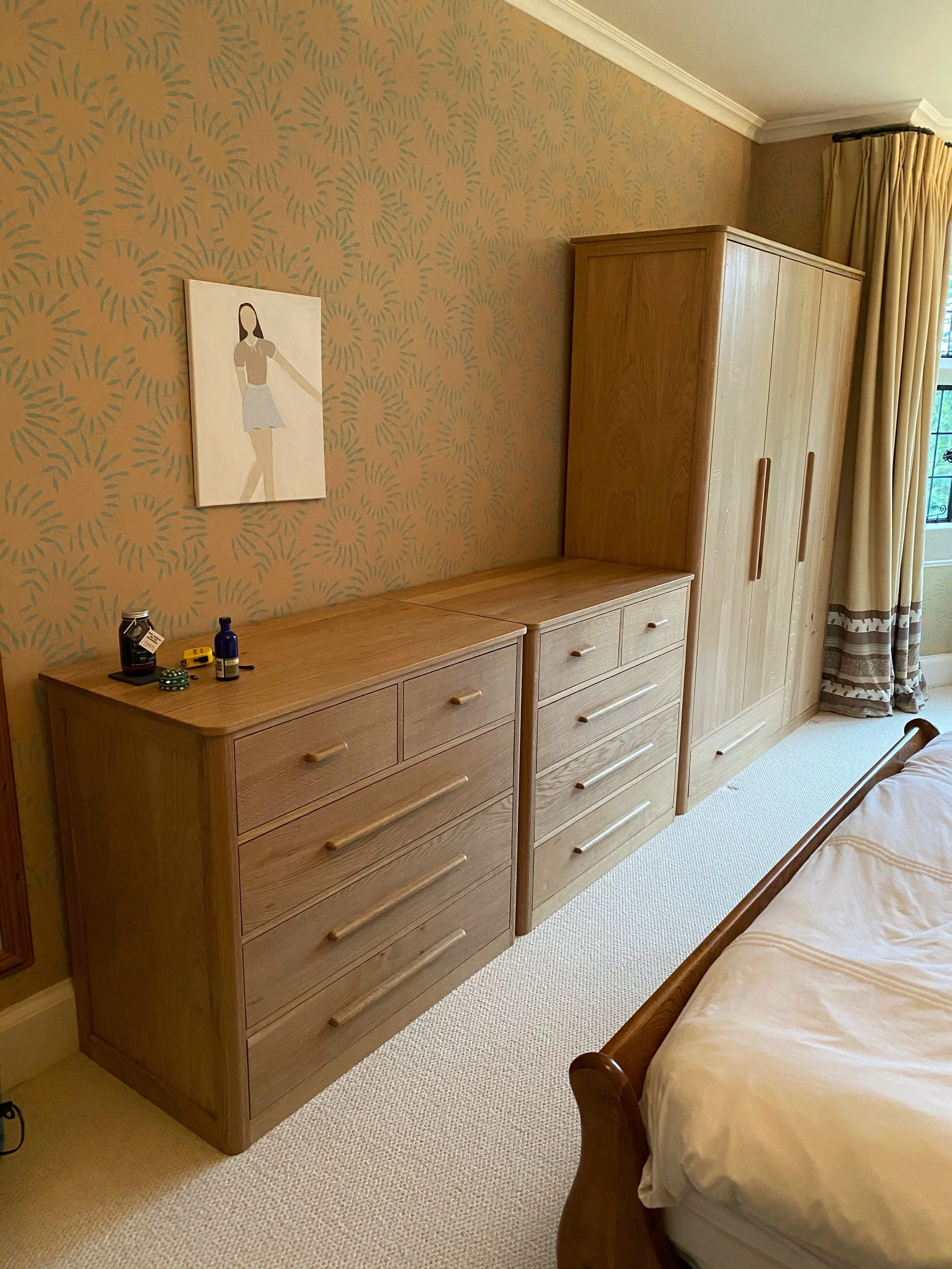 A wooden dresser with six drawers sitting beside a bed, against a wallpapered wall with a floral pattern. On top of the dresser are small objects including bottles, a tape measure, and a small container. To the right, there is a wooden wardrobe next 