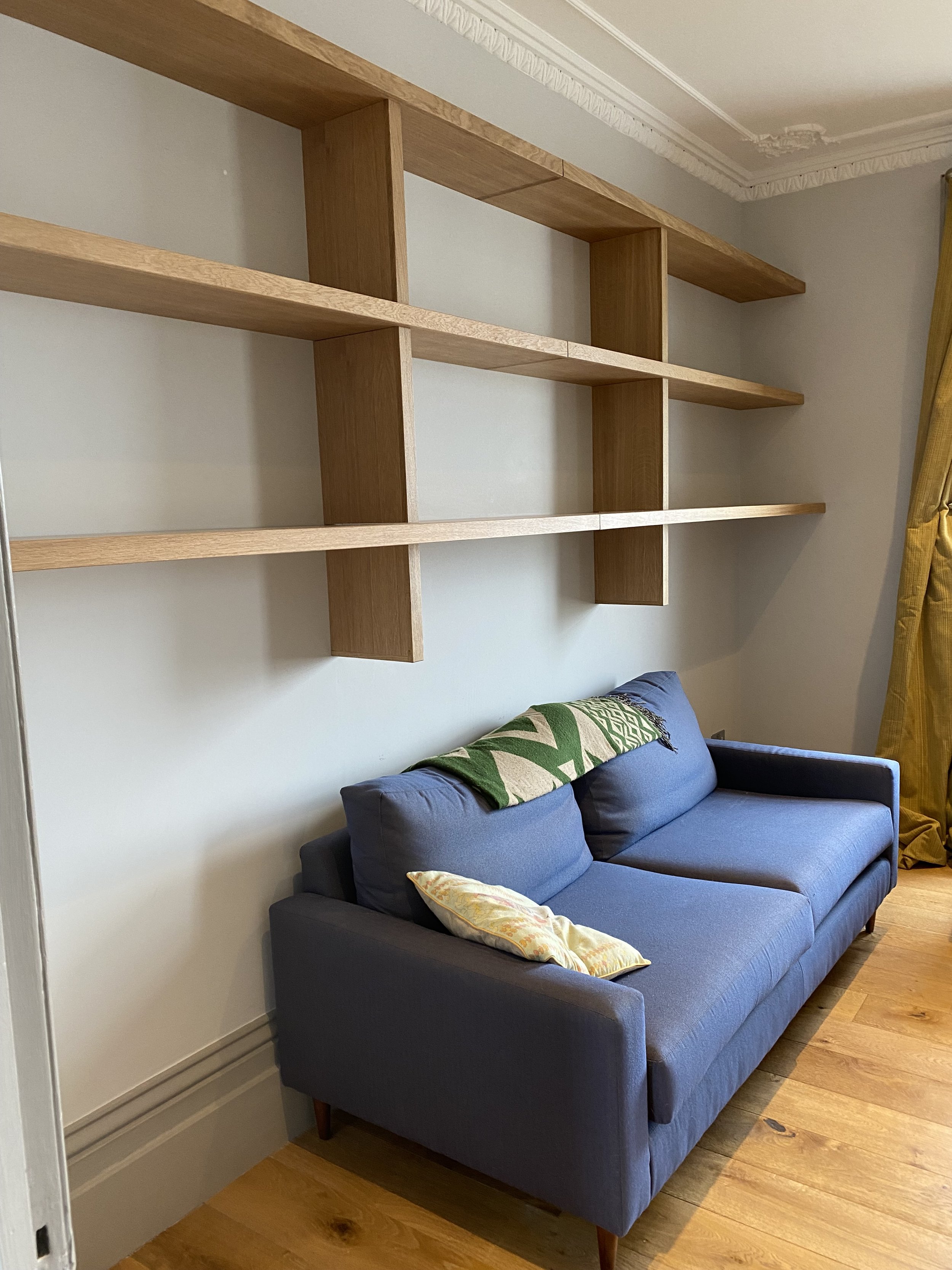 A blue sofa with two pillows, one yellow and one patterned, in front of a light-colored wall with a wooden bookshelf mounted above it. There is a yellow curtain on the right side of the image and wooden flooring.