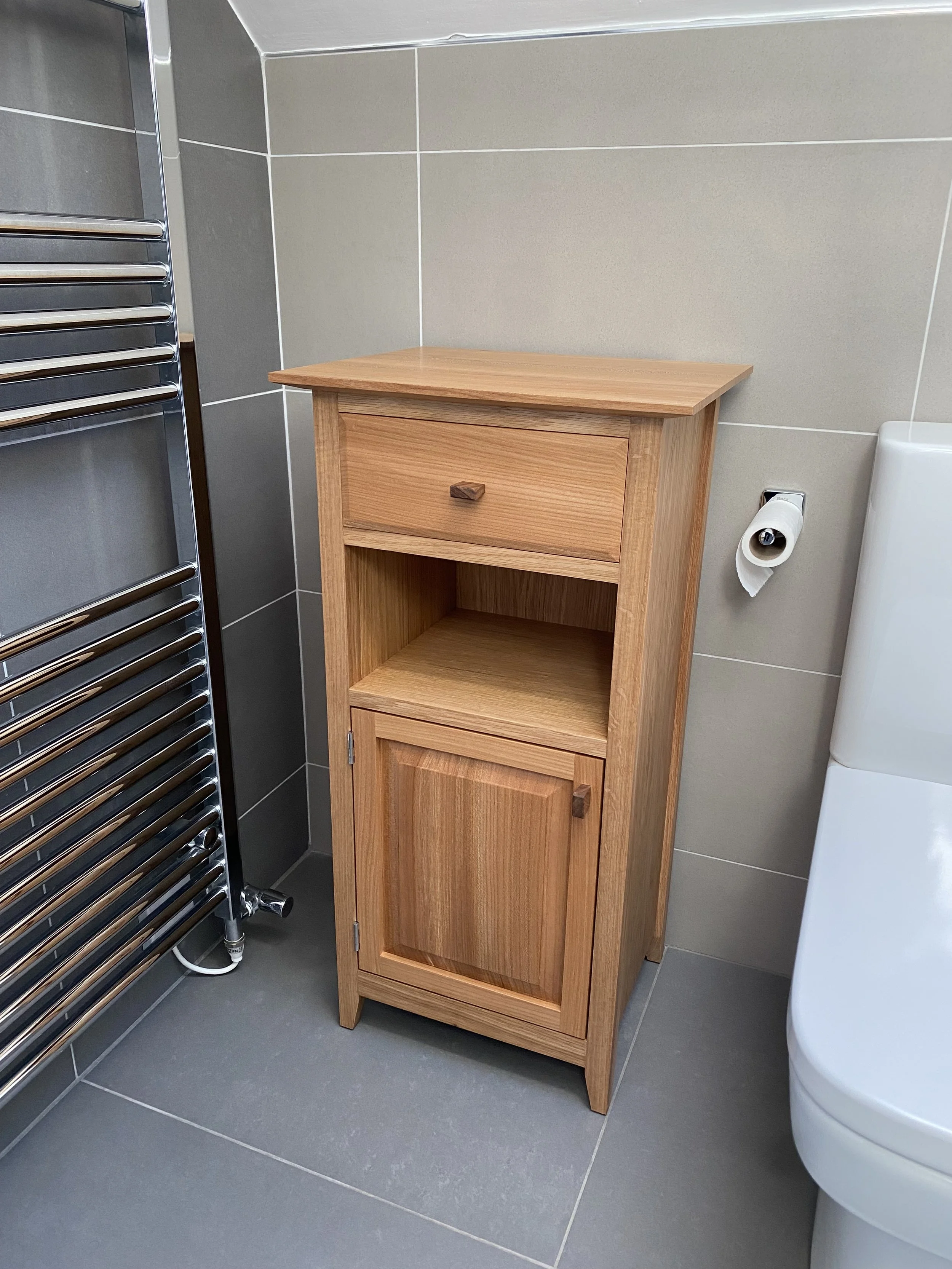 A small wooden bathroom cabinet with a drawer and an open shelf, placed beside a modern toilet and a heated towel rail in a bathroom with gray tiled walls and floor.