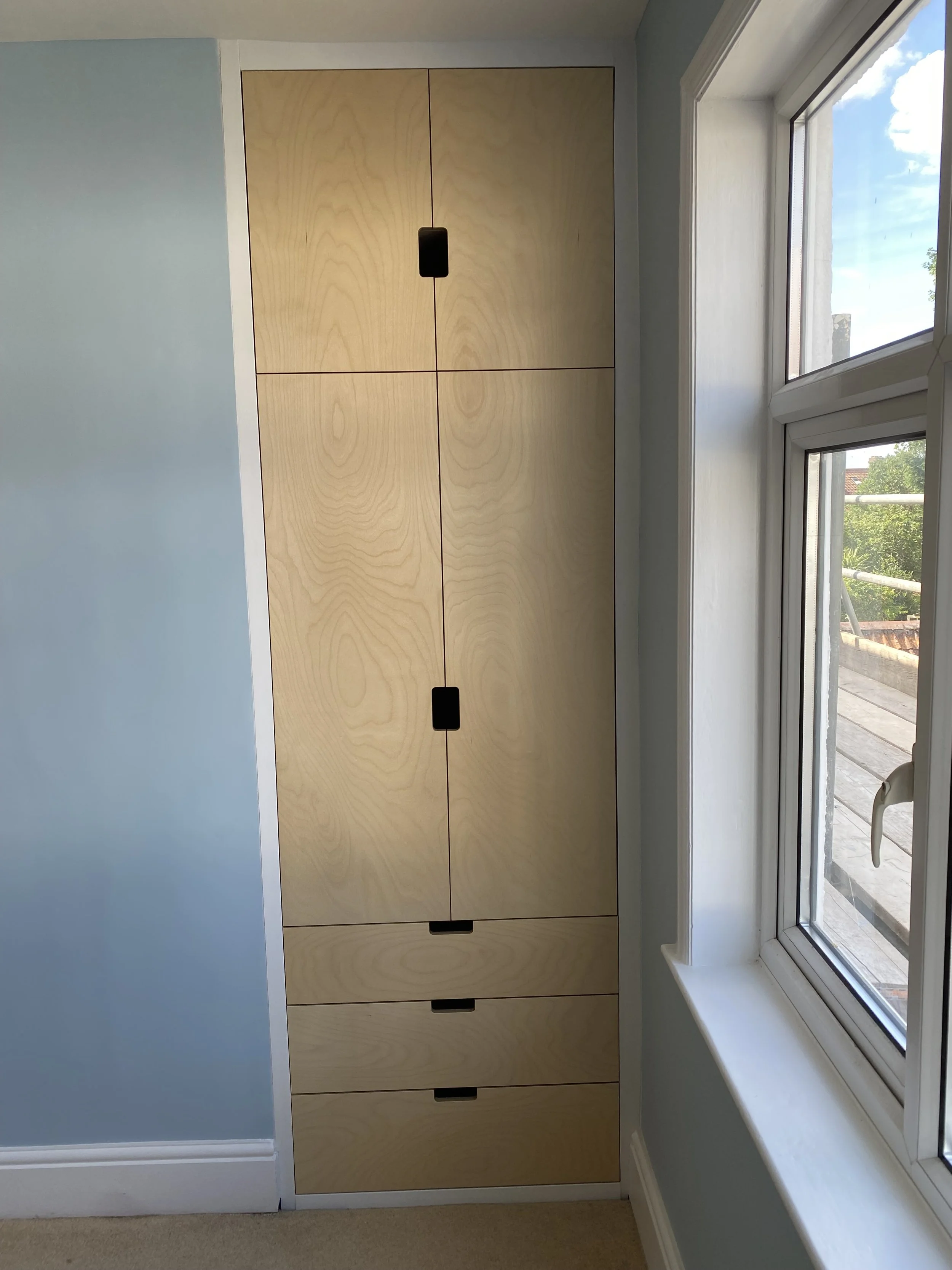 Wooden cabinet with four doors and black handles next to a white-framed window showing a clear sky and part of a balcony with railing.