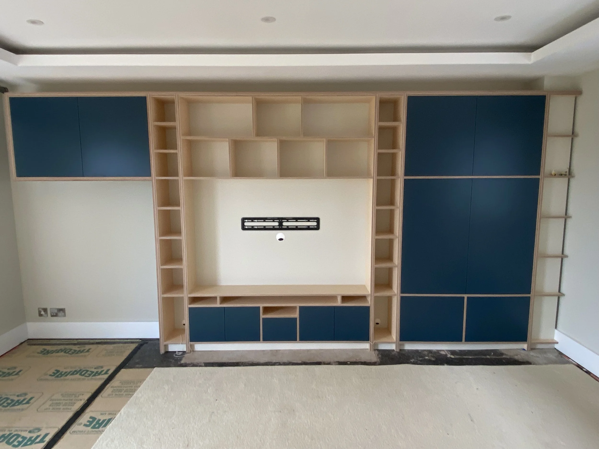 Empty living room with built-in wooden shelves and cabinets in progress, with some blue doors, and under construction floor covering.