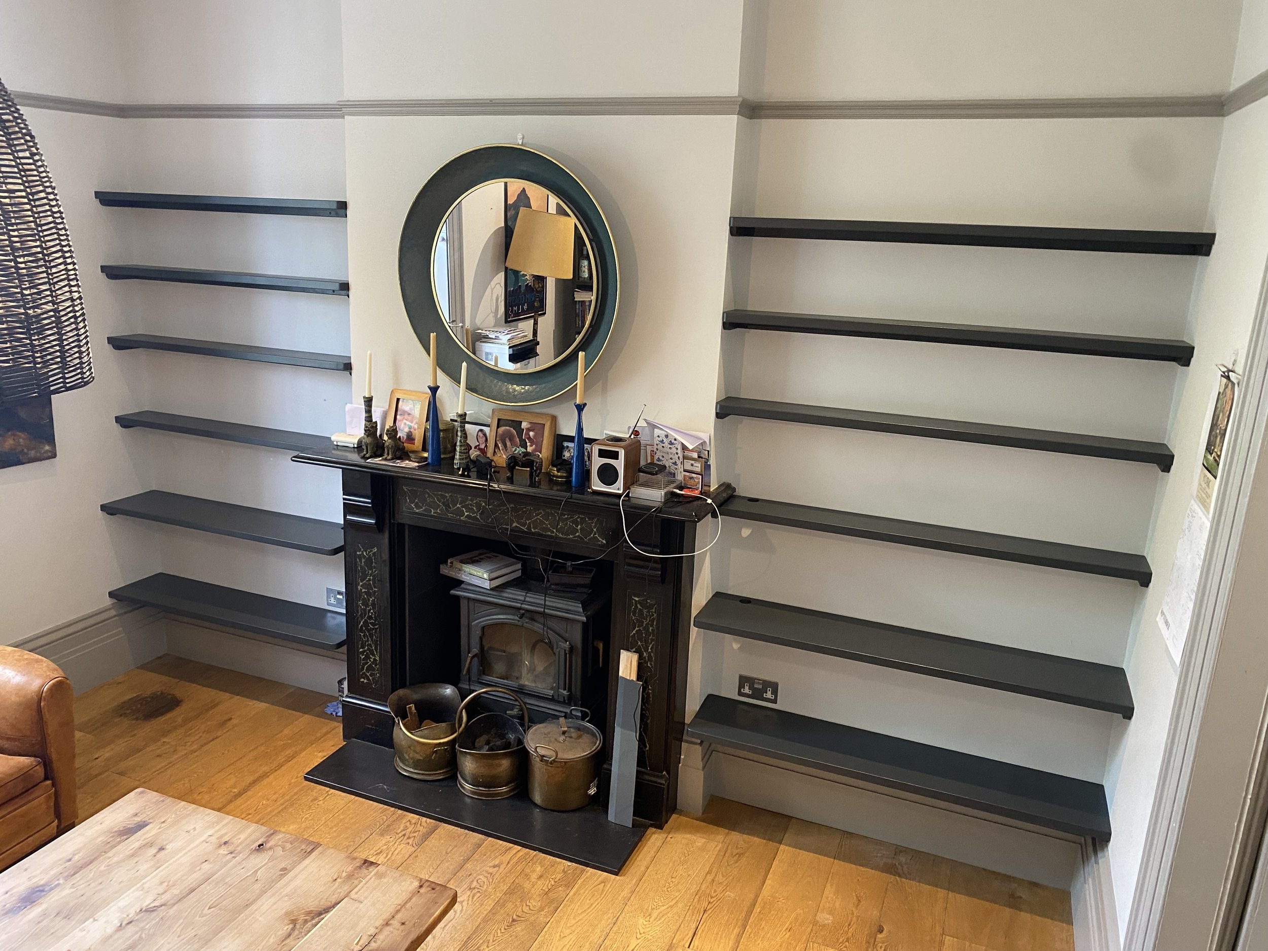 A living room corner with empty black floating shelves on white walls, a black fireplace, a mirror, and various decorative items and framed photos on the mantel.
