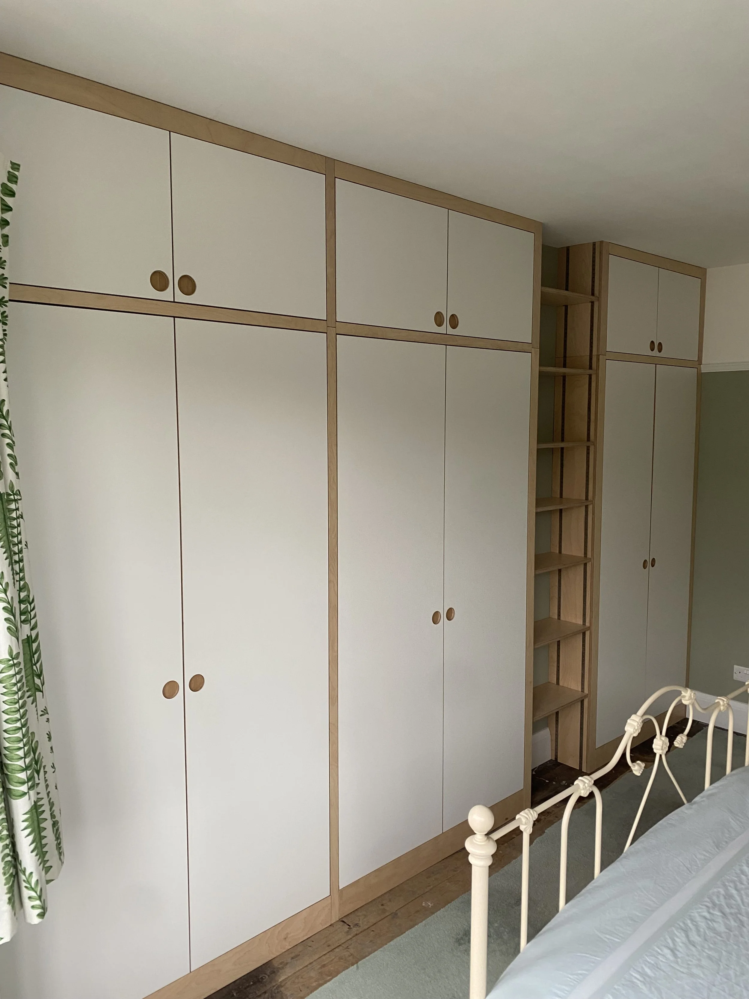 A bedroom with white wooden wardrobes with round handles, a white metal bed frame, green and white patterned curtains, and a wooden floor.
