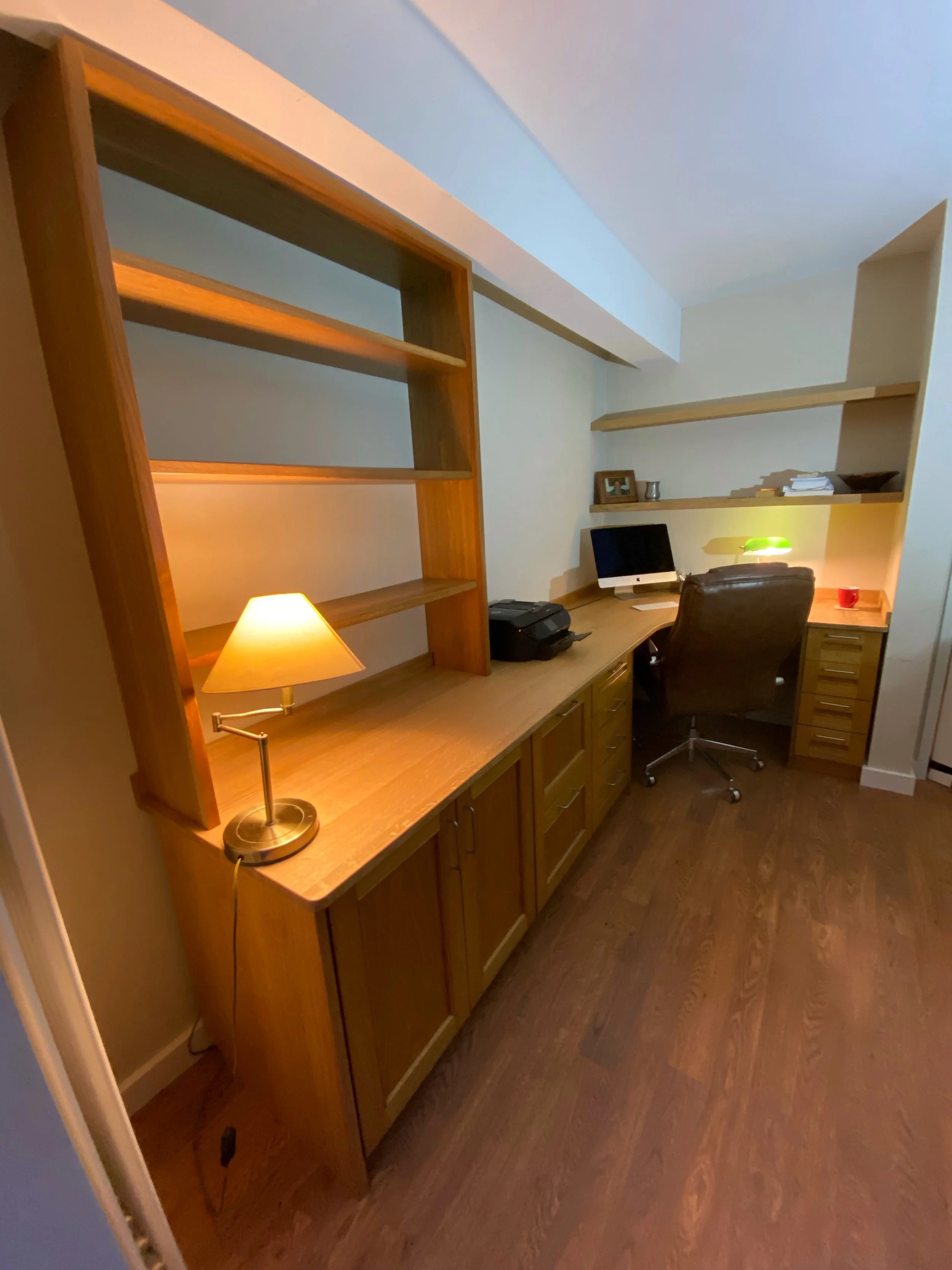 Home office with wooden desk, black computer, table lamp, shelves with books and decor, and wooden flooring.
