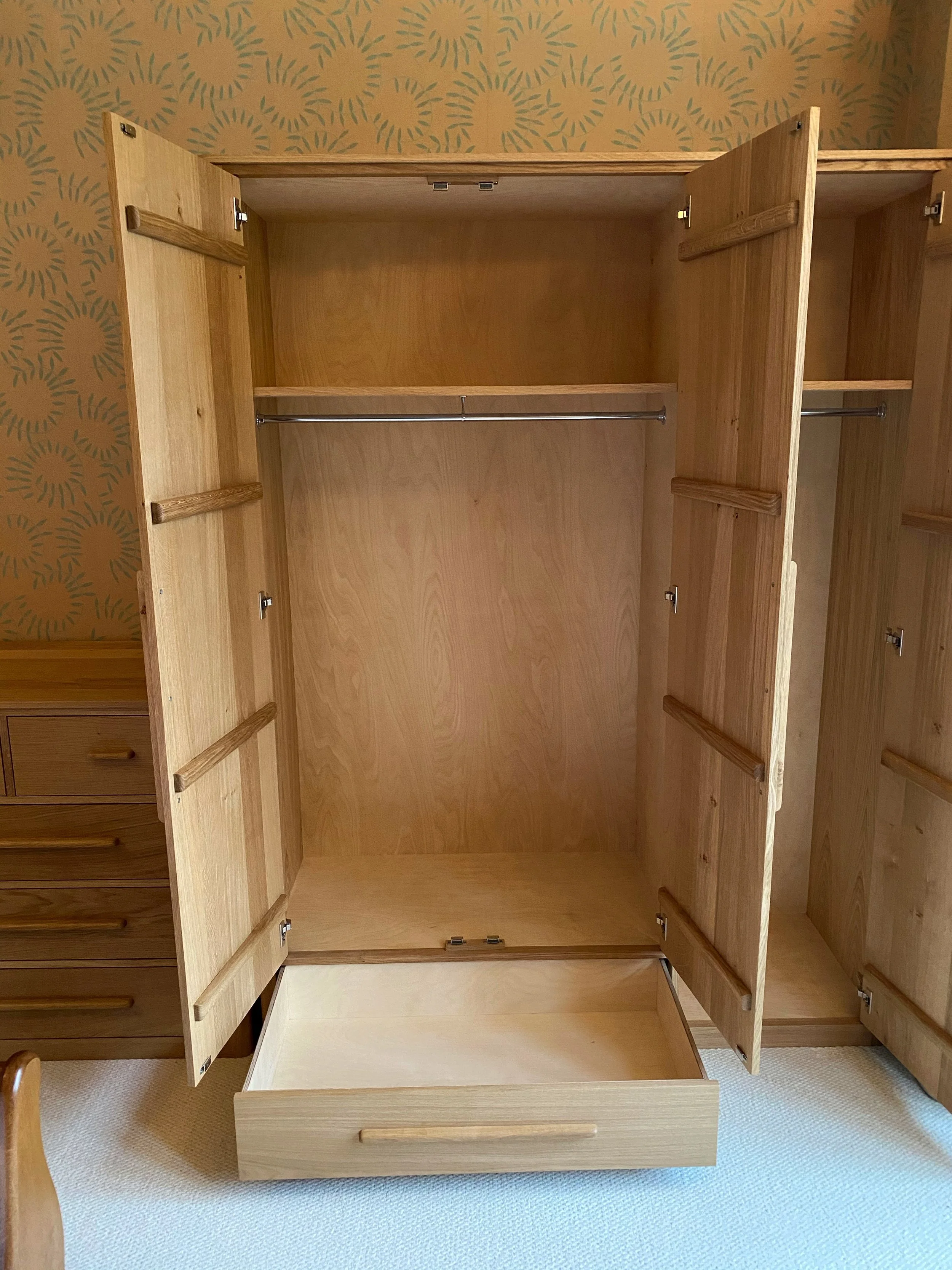 Empty wooden wardrobe with open doors showing hanging rod inside, positioned against a wall with patterned wallpaper.
