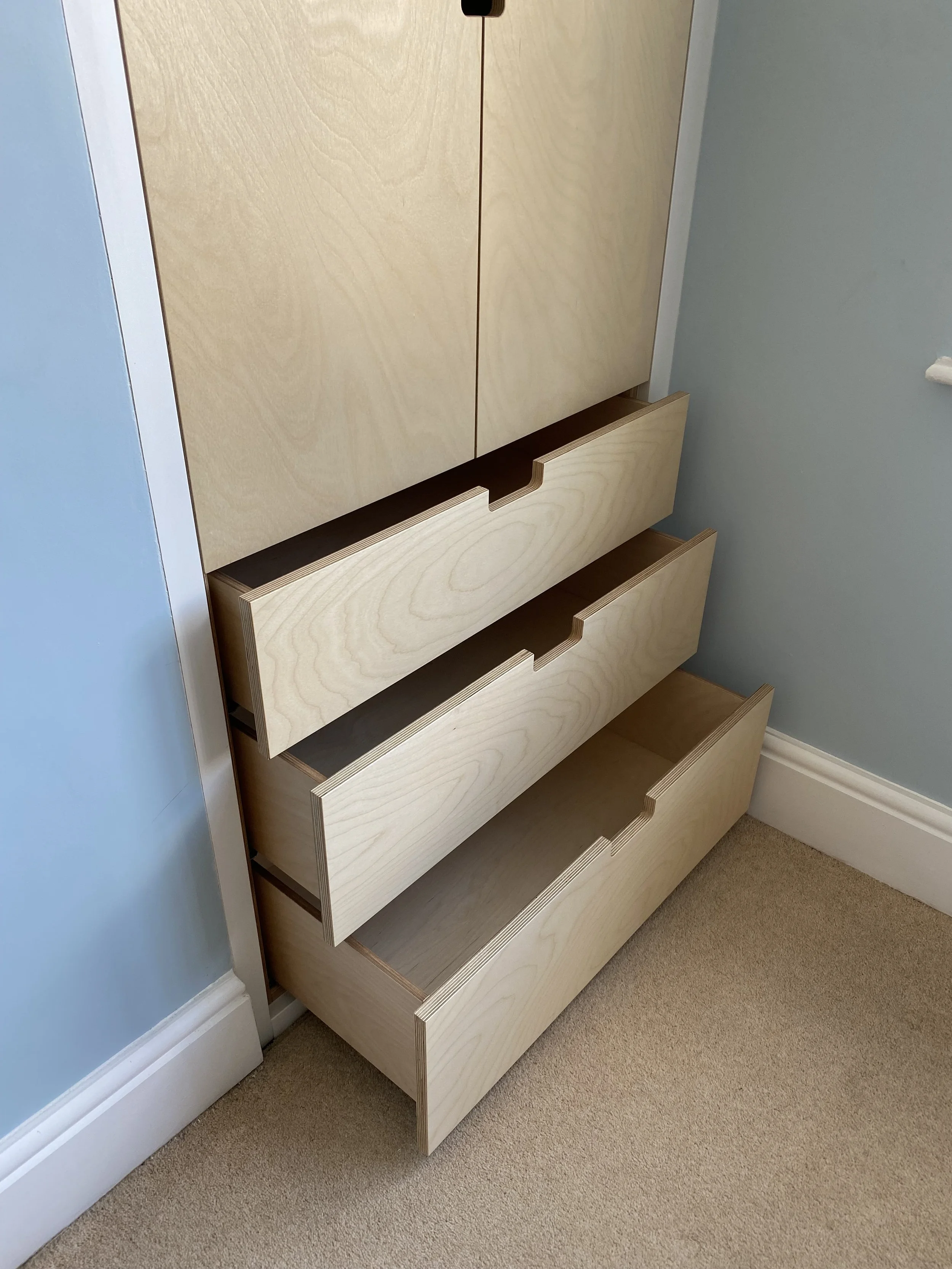 Multiple beige wooden drawers partially pulled out of a tall cabinet in a room with beige carpet and light blue walls.
