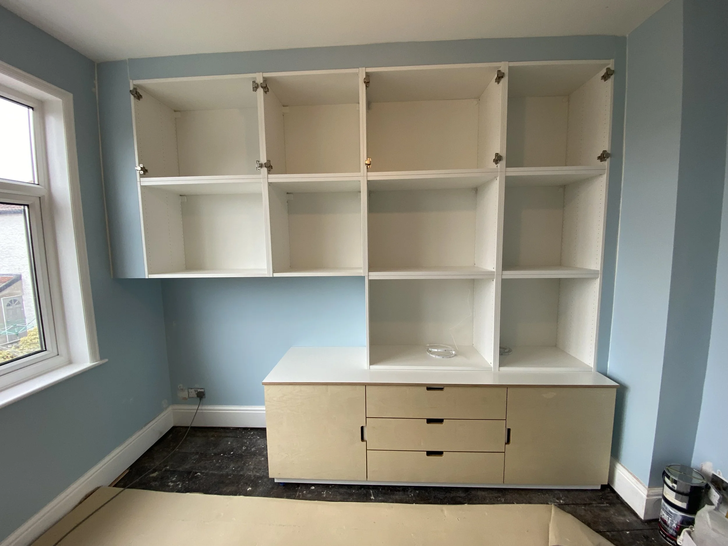 Unfinished white wall cabinet with drawers below in a room with blue walls and a window.