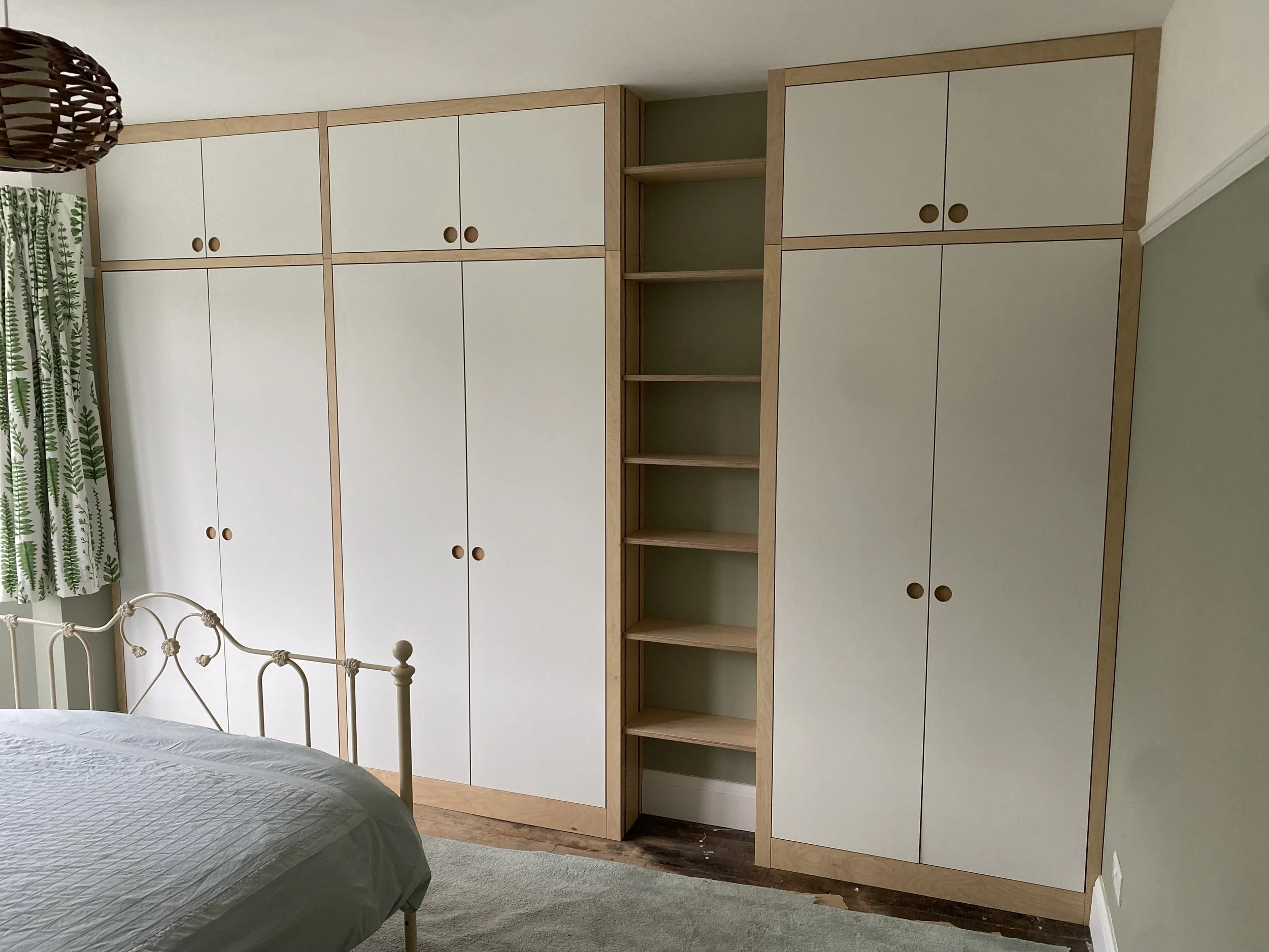 Bedroom with white and wood closet doors and open shelving unit, part of a bed with a gray or green blanket, and a window with green leaf patterned curtains.