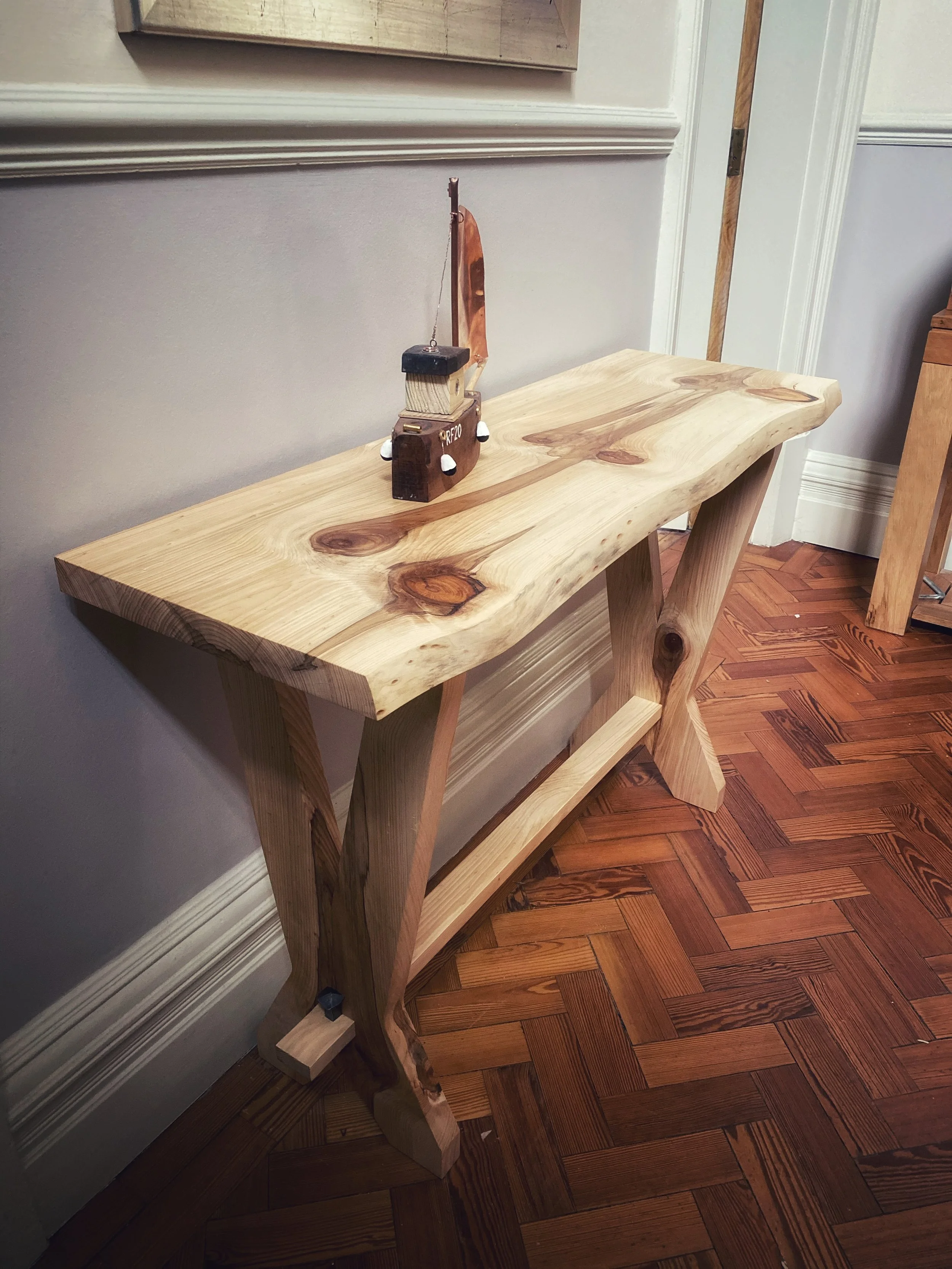 Wooden table or bench with a wooden model ship on it, placed against a wall near a door with hardwood flooring.
