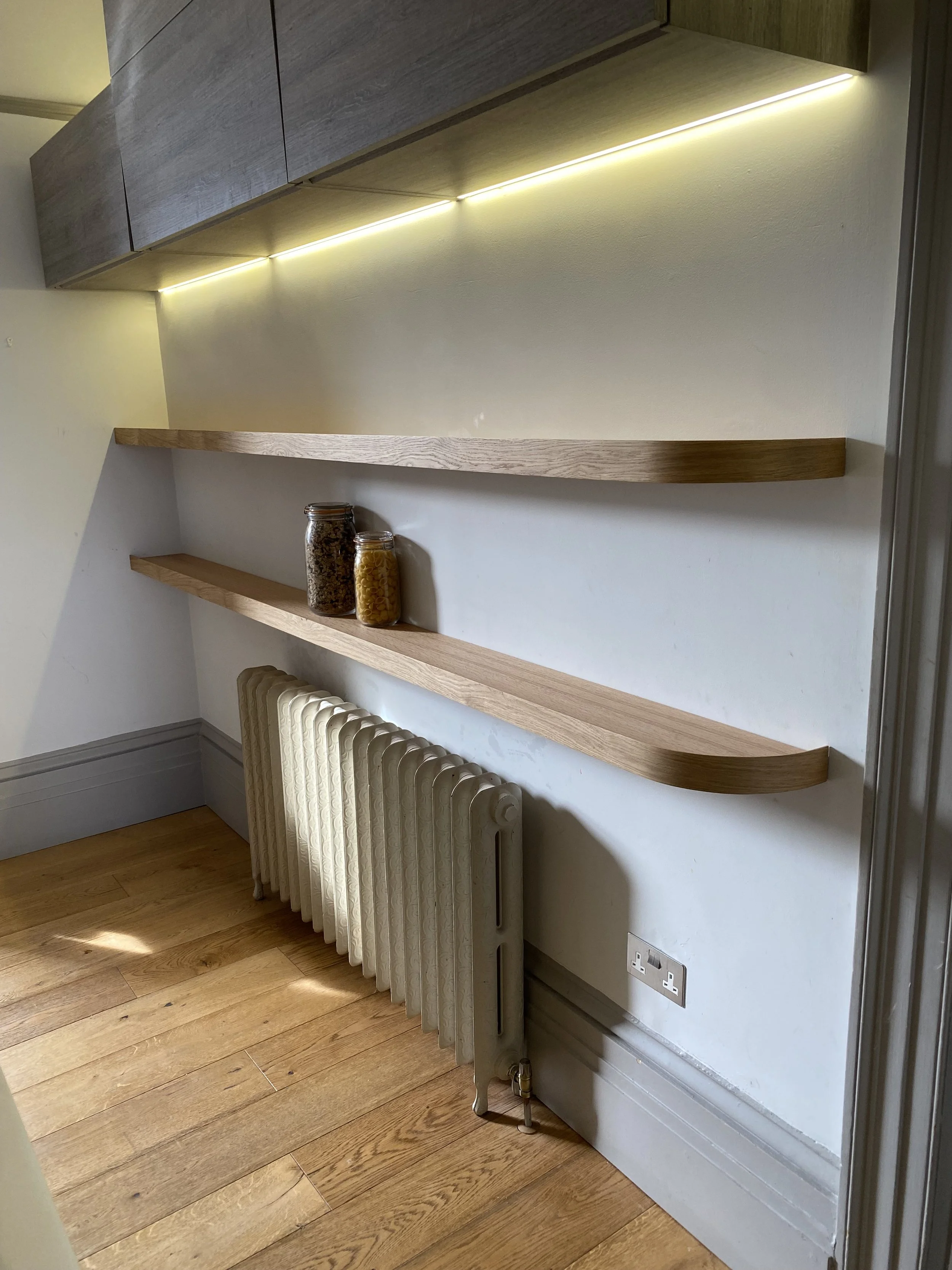 Interior corner with a white radiator heater, wooden shelves, and two glass jars, one filled with pasta and the other with cereal, a wall socket, and a wooden floor.